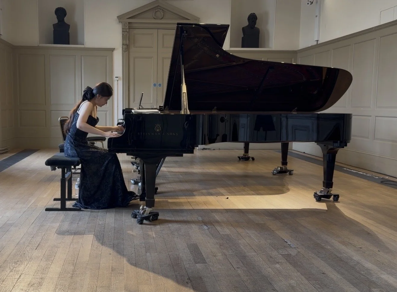 A woman in a dark dress playing a grand piano in a spacious room with wooden floors and wood-paneled walls.