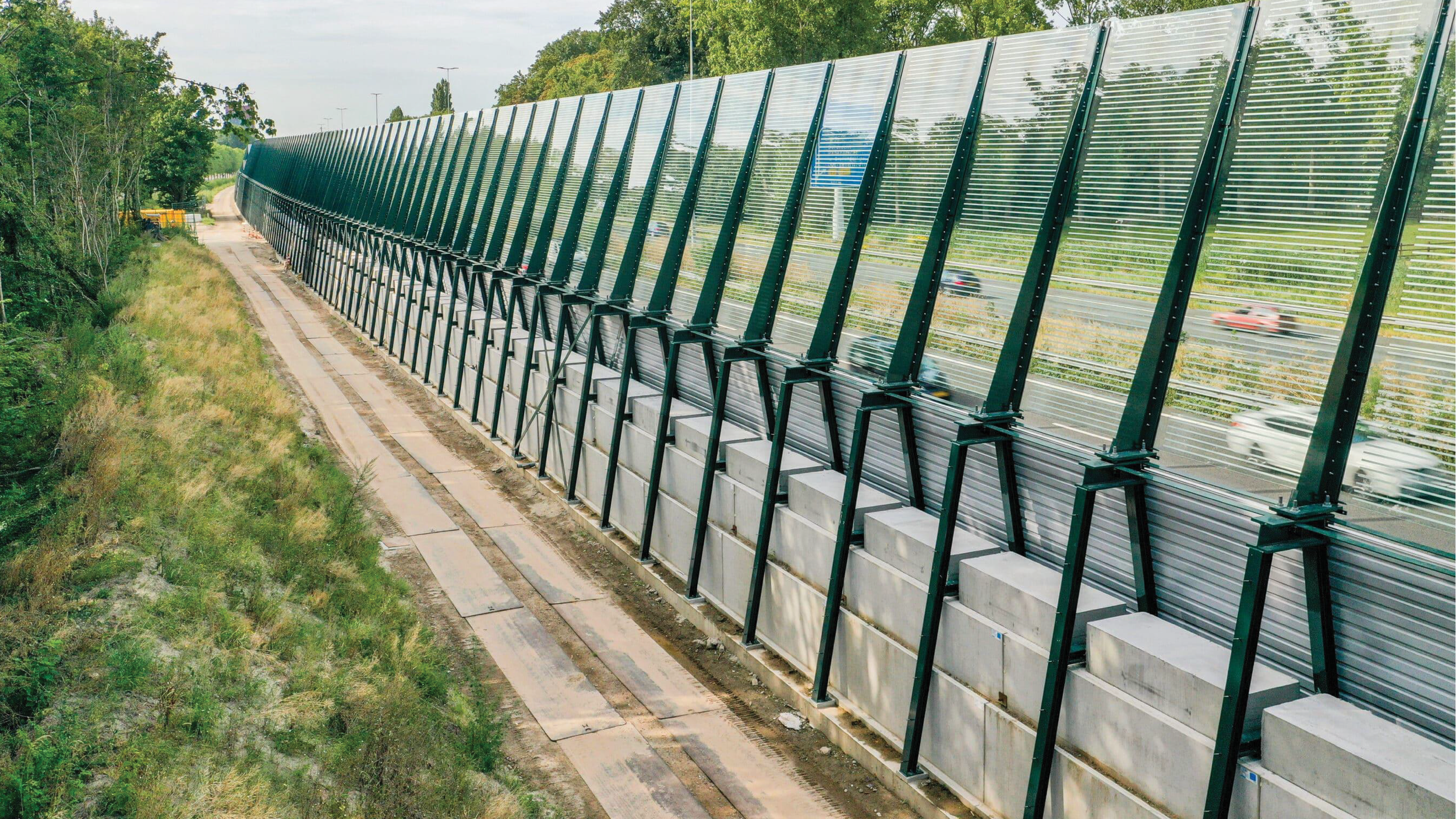 Highway sound barrier with green metal panels and supporting structures, alongside a road with cars visible through the panels, surrounded by green trees and grass, under a partly cloudy sky.