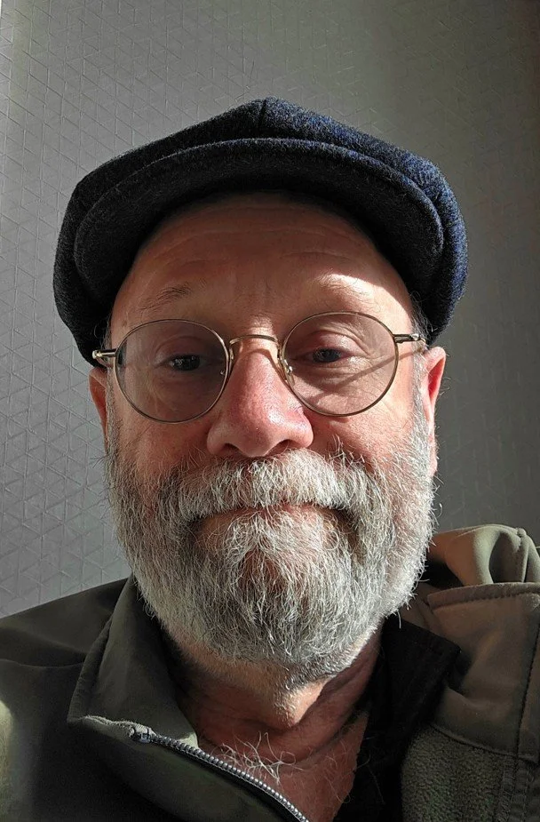 Close-up of a smiling older man with a gray beard, round glasses, wearing a dark cap, and a zip-up jacket.