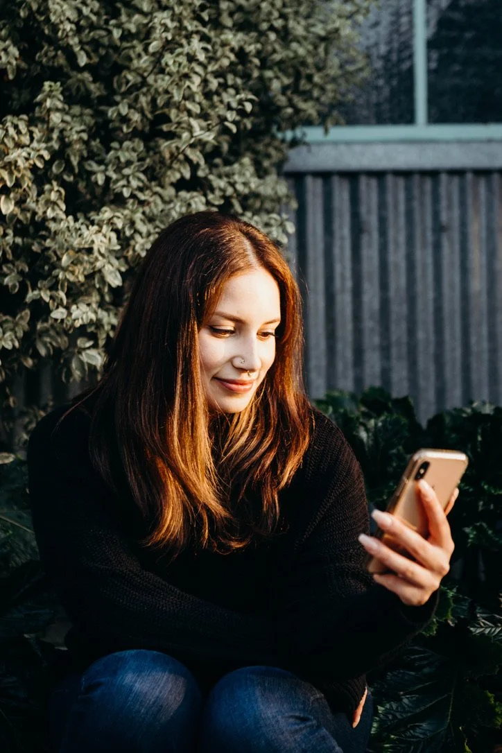 A young woman with long brown hair and light skin sitting outdoors, smiling as she looks at her smartphone, surrounded by greenery and a wooden fence.