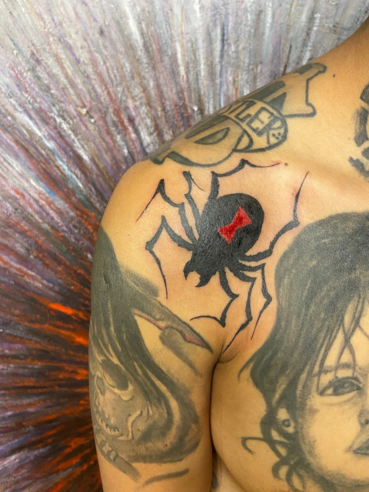 Close-up of a person's shoulder and upper arm with a tattoo of a spider, a heart with a cross, and a cartoonish face, against a colorful textured background.