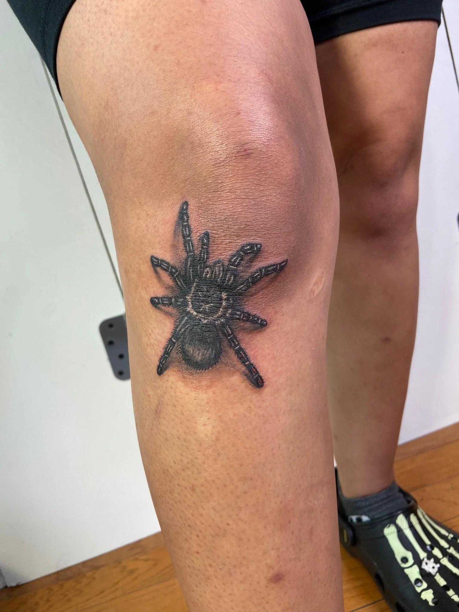 Close-up of a person's upper thigh with a tattoo of a spider and part of a black sneaker with yellow design visible in the background.