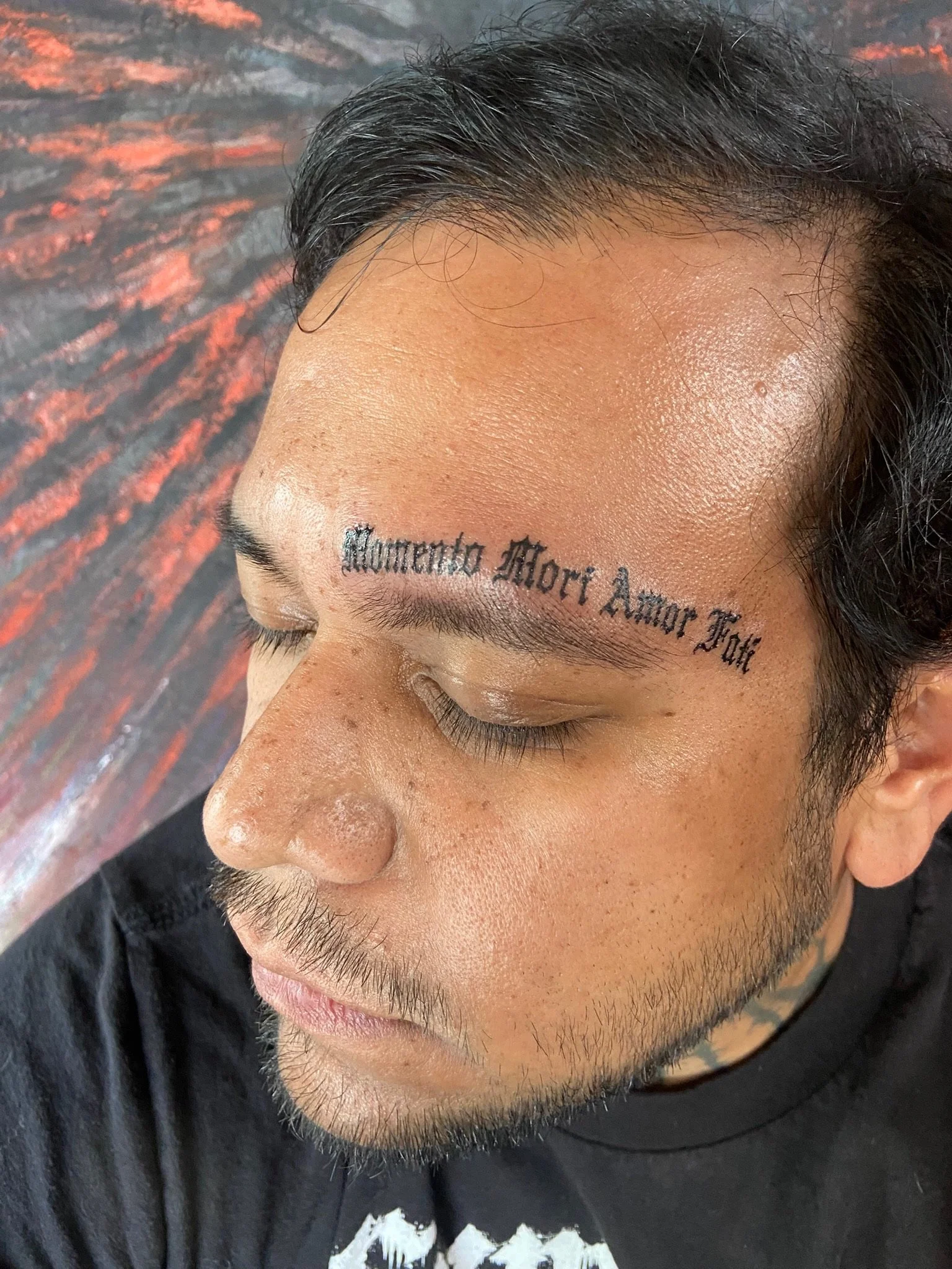Close-up of a person's forehead with a tattoo in cursive script that reads 'Momento Morí Amor Tua', with dark hair and a mustache, on a background of a dark, textured surface.