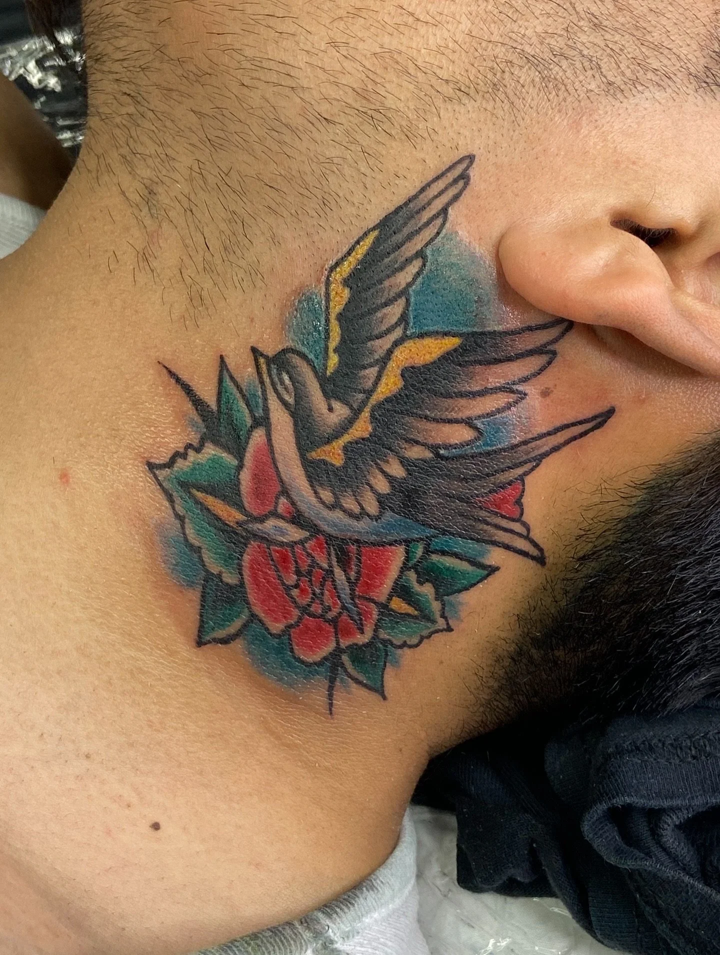 Tattoo of a bird with outstretched wings and a heart on a person's neck, with colorful shading and floral elements.