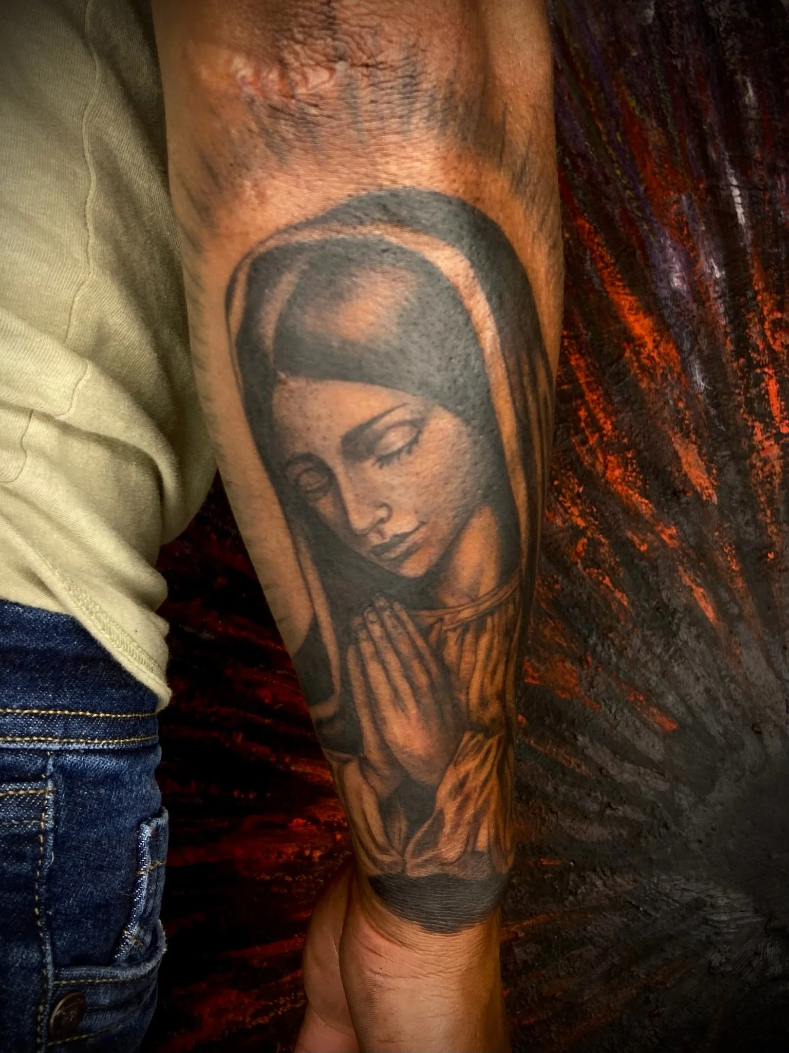 Close-up of a tattooed person's arm featuring a portrait of a woman with closed eyes and long eyelashes, with a dark background and orange and red lighting.