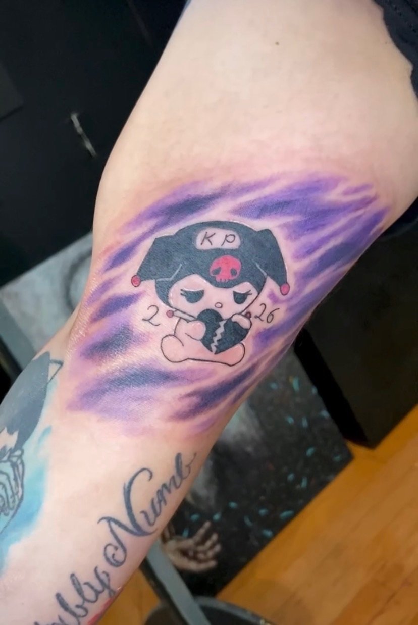 Tattoo of a chibi girl with a broken heart, holding a phone, with the numbers '2026' and the letters 'KP' above her head. The background features purple and blue swirling colors.