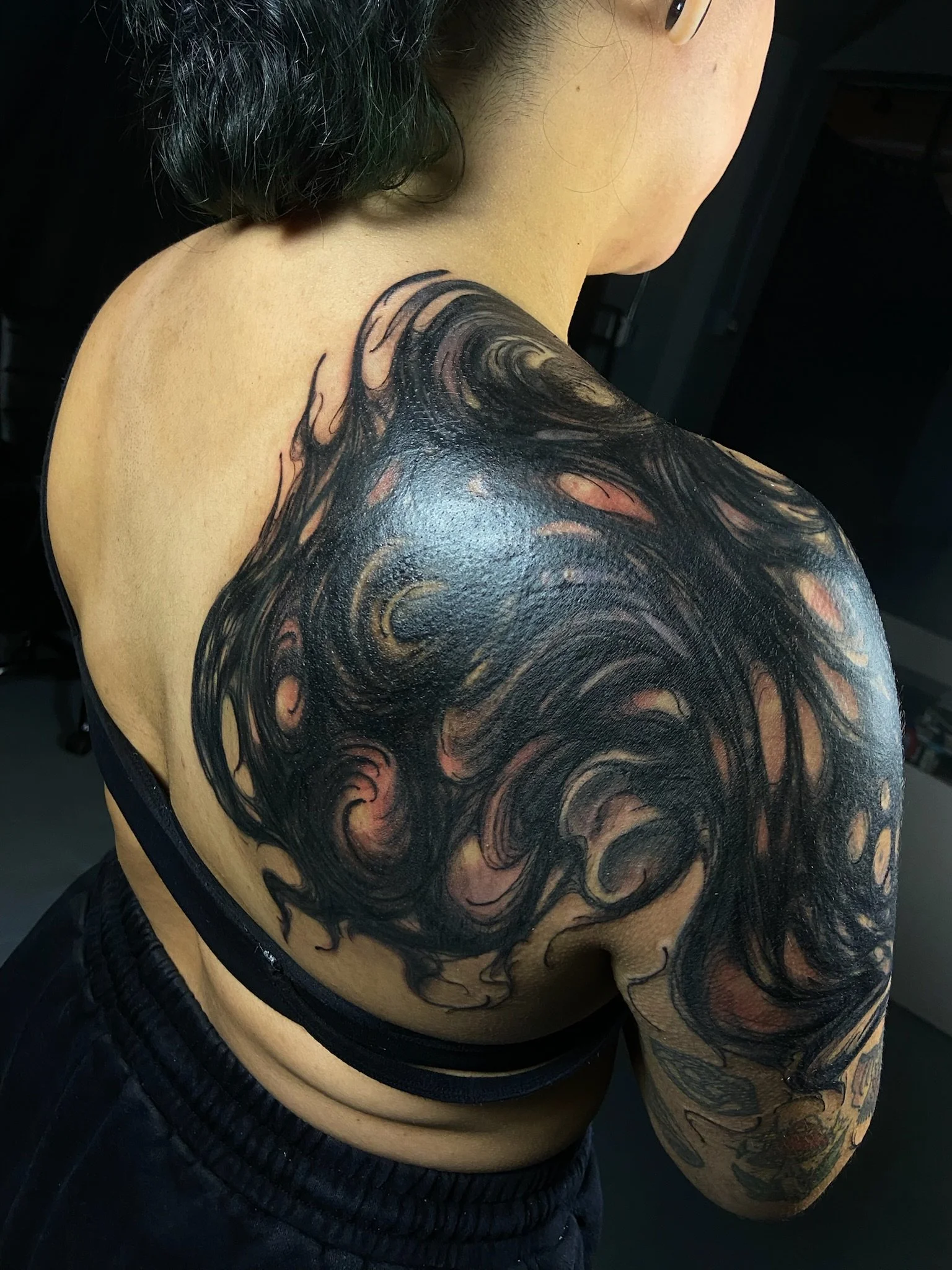Close-up of a person's shoulder and arm with blackwork swirl coverup large-scale shoulder tattoo, wearing a black shirt and black shorts.