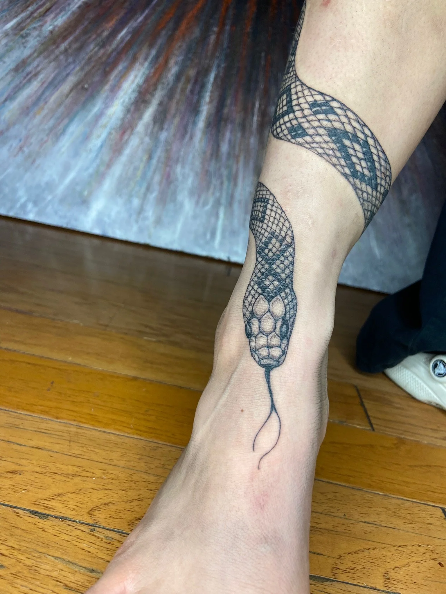 A person's leg with a large black snake tattoo running from the ankle to above the knee. The tattoo depicts a snake with a patterned body and a heart-shaped head, with a tail extending downward. The leg is resting on a wooden floor near what appears 