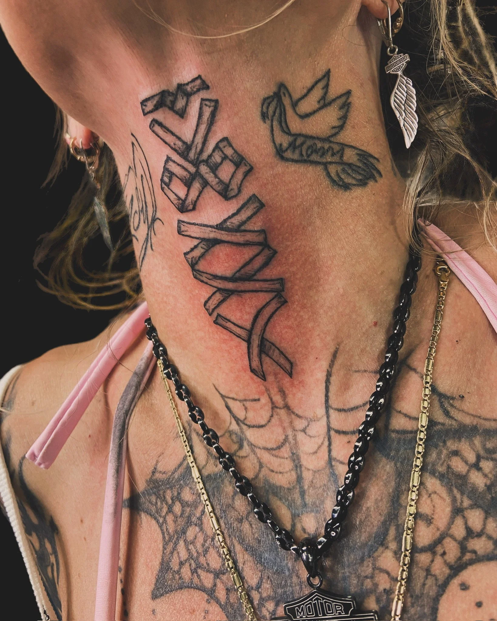 Close-up of a person's chest with multiple tattoos, including a large spider web, a bird with a banner reading 'Mom,' and a stylized word tattoo. The person is wearing several necklaces and a Harley-Davidson badge.