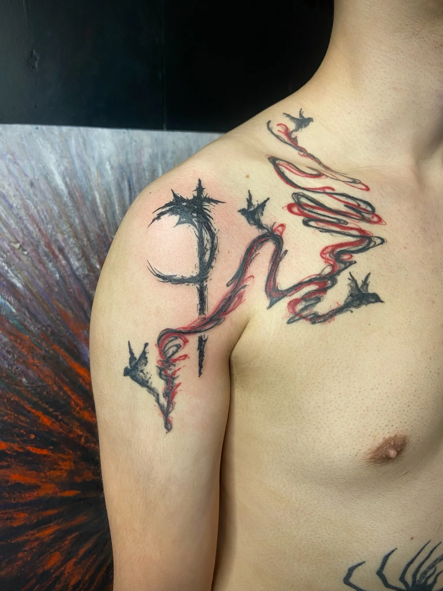 Close-up of a person's shoulder and chest with a tattoo of a line of text in red and black ink, with a background of a dark wall and textured surface.