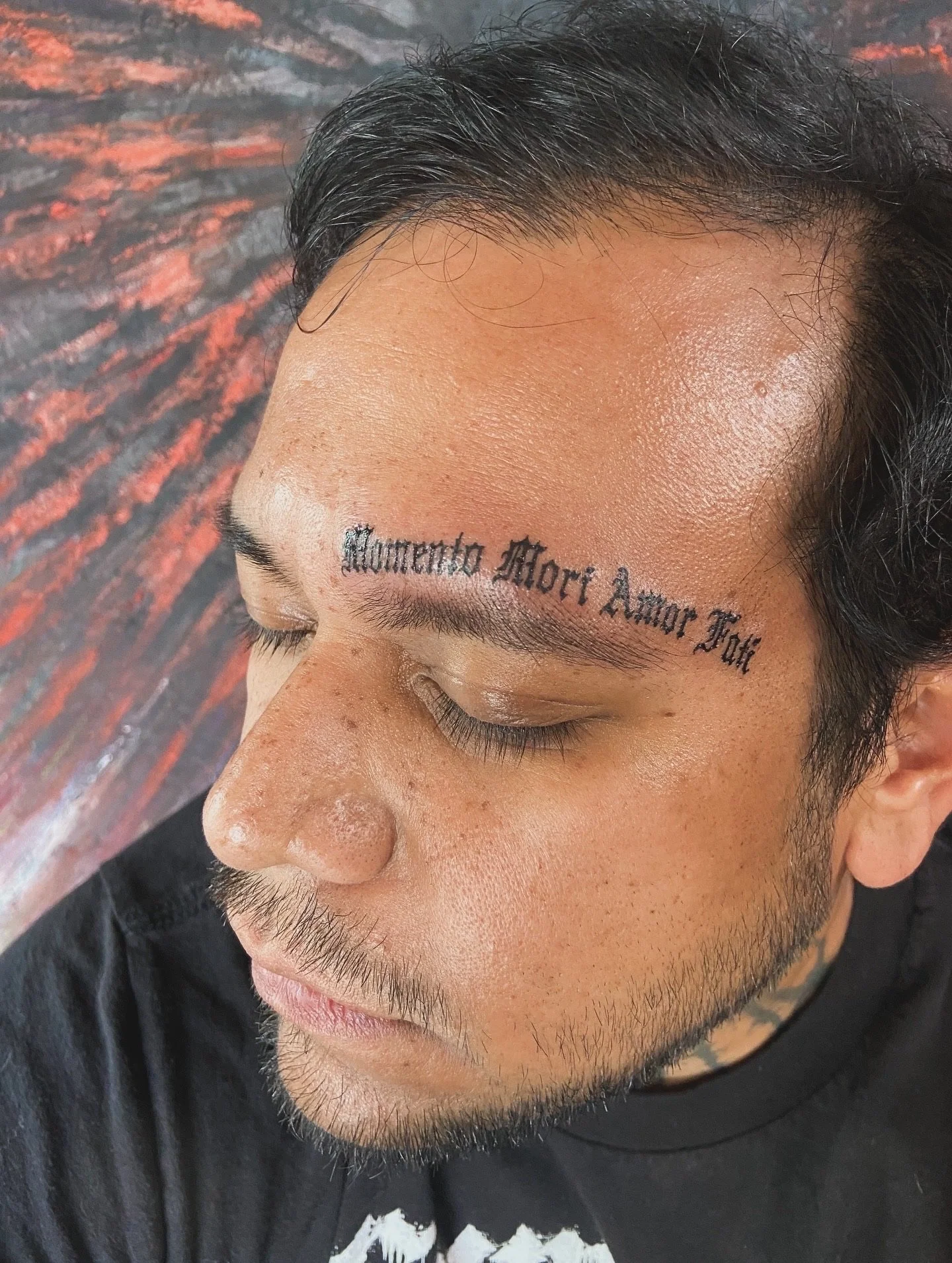 Close-up of a man's face with a tattoo above his right eyebrow that reads 'Momento Morí Amor Fati' in black ink.