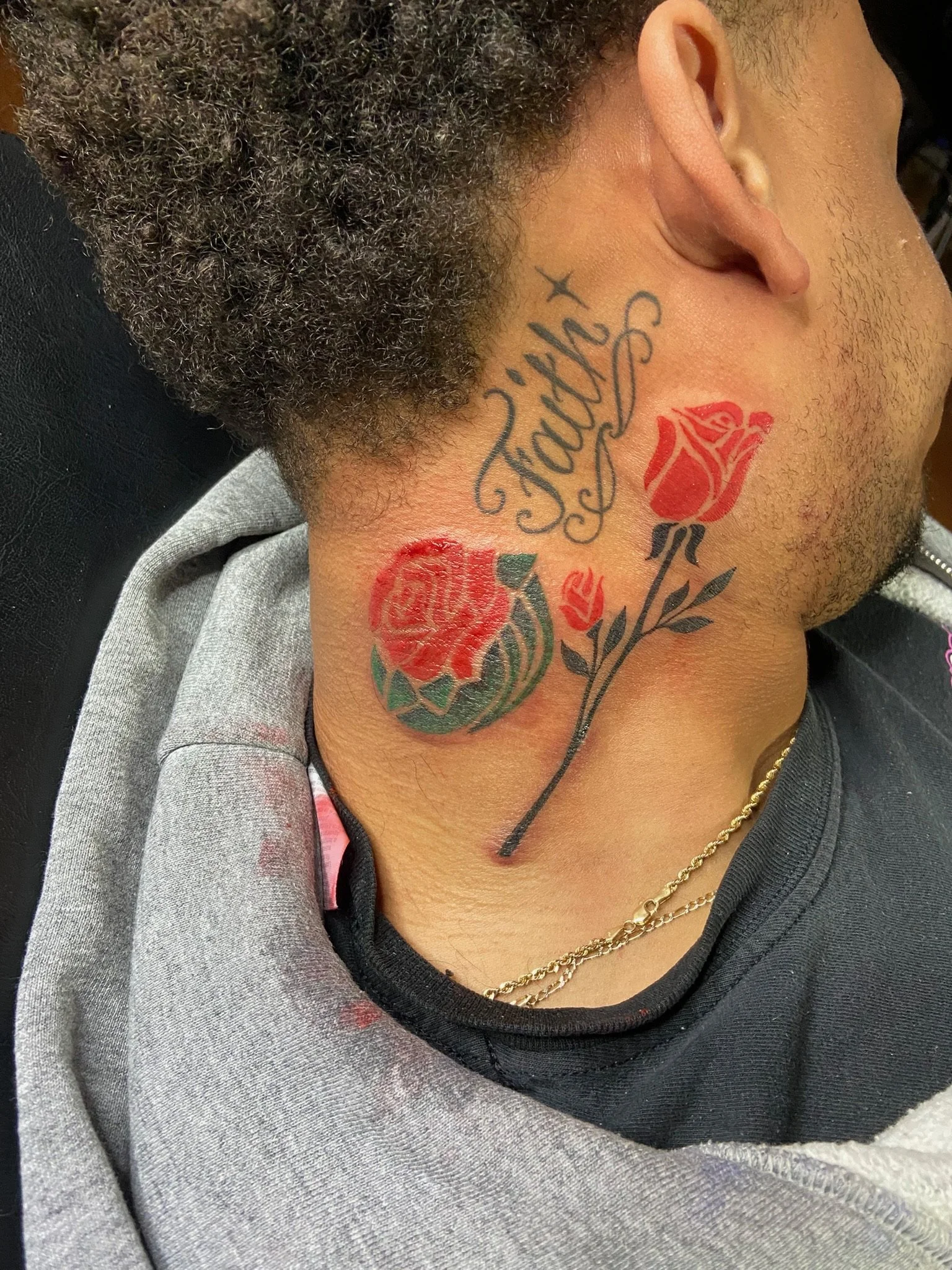 Close-up of a person's neck showing tattoos of the word 'Family' and a red rose with green leaves, with part of their face, ear, and beard visible, wearing a black and gray hoodie and a gold chain.