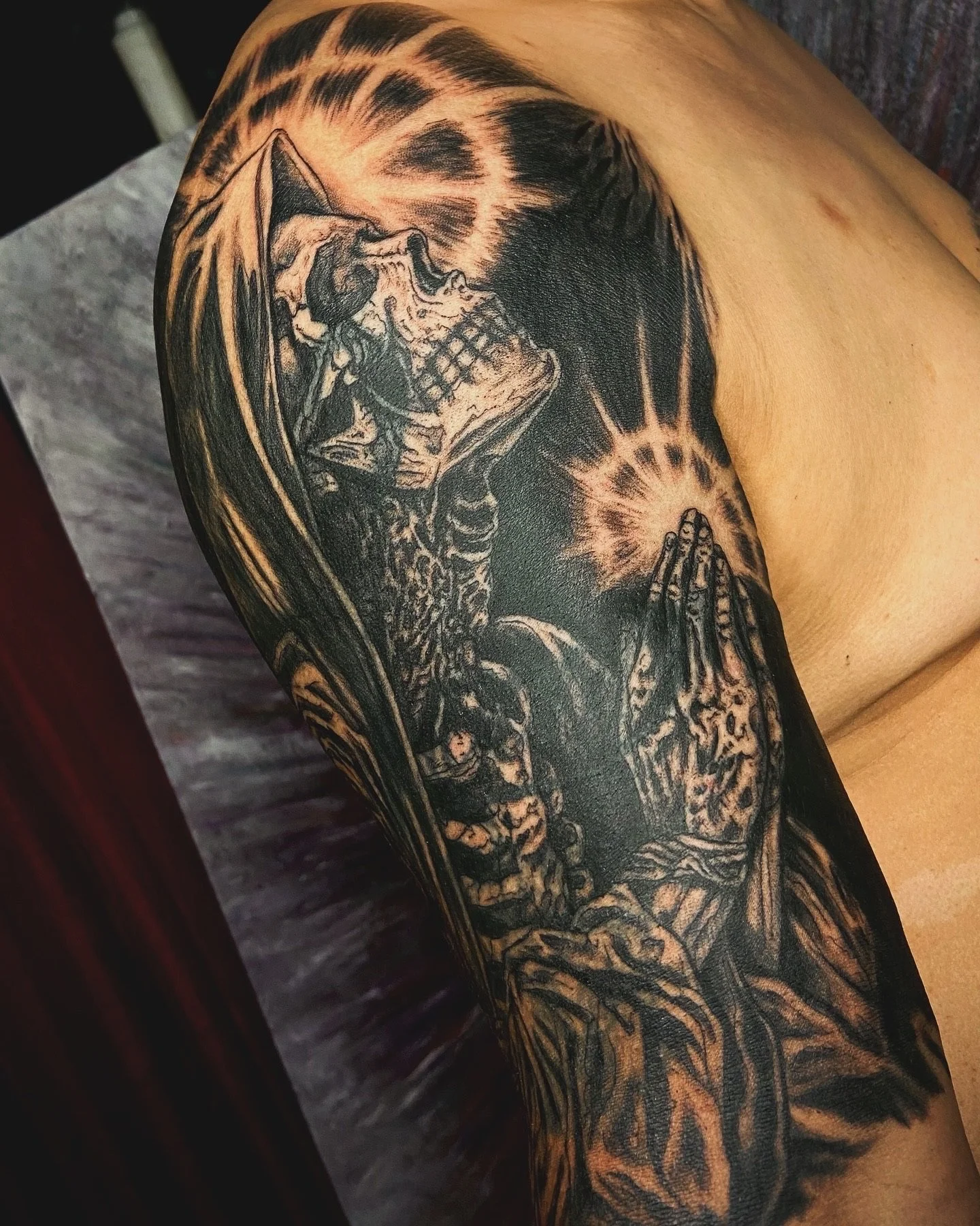 Black and gray tattoo of a human skull, hands in prayer, and angel wings on arm.