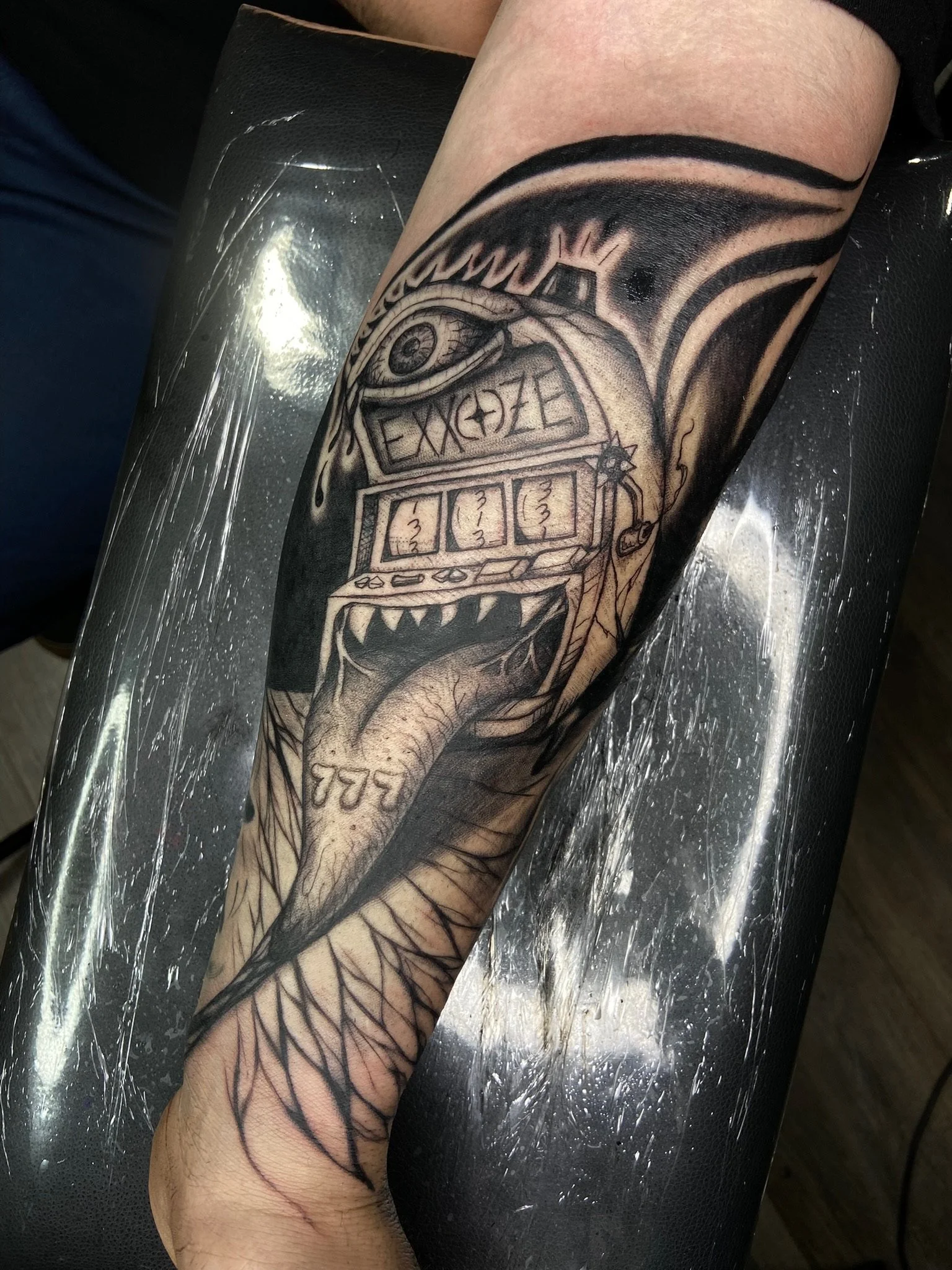 A tattoo on a person's forearm depicting a detailed eye at the top, with a slot machine beneath it showing three sevens, and various abstract and line work elements surrounding the design.