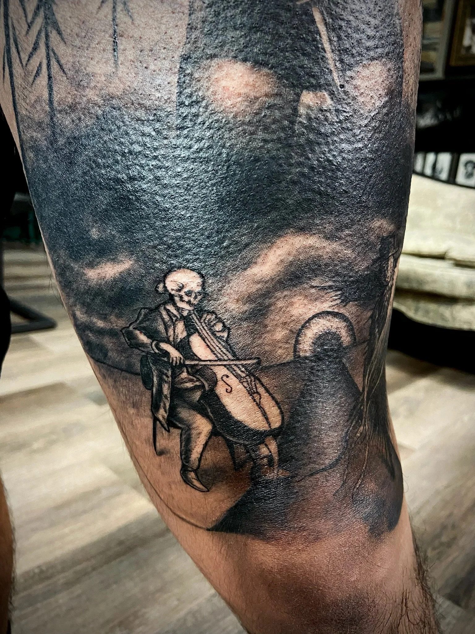 Close-up of a black and gray tattoo on a person's upper arm, featuring a skeleton and a sword.