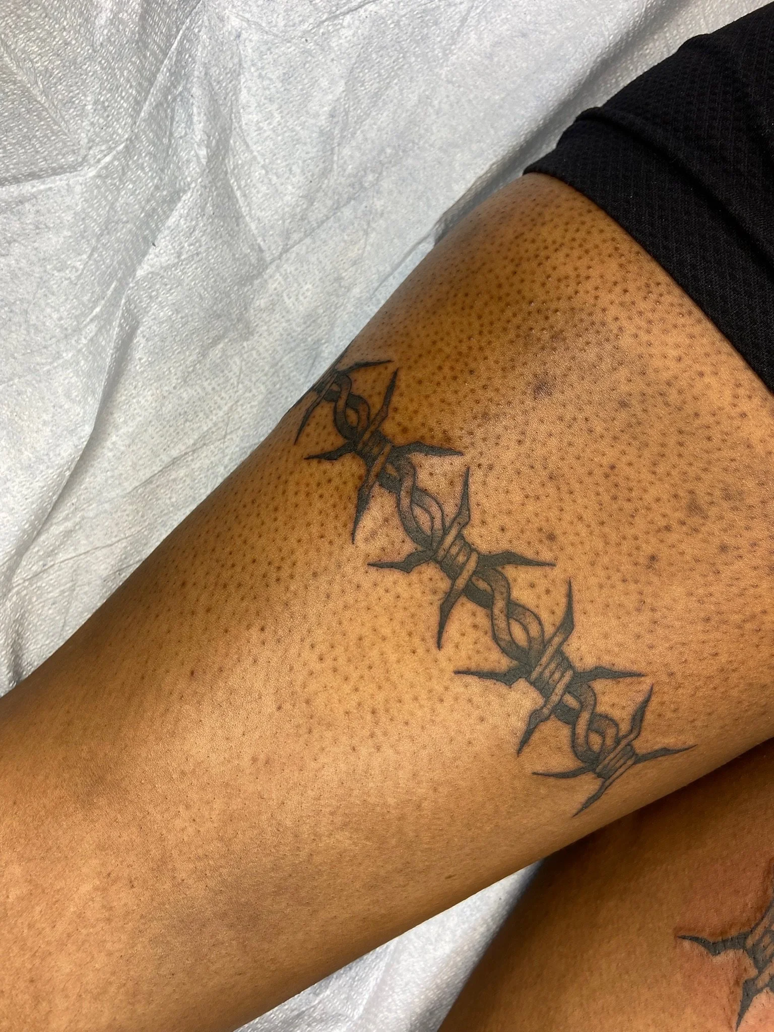 Close-up of a person's thigh with a tattoo of a barbed wire design, wearing black fishnet tights and black shorts, against a white textured surface.