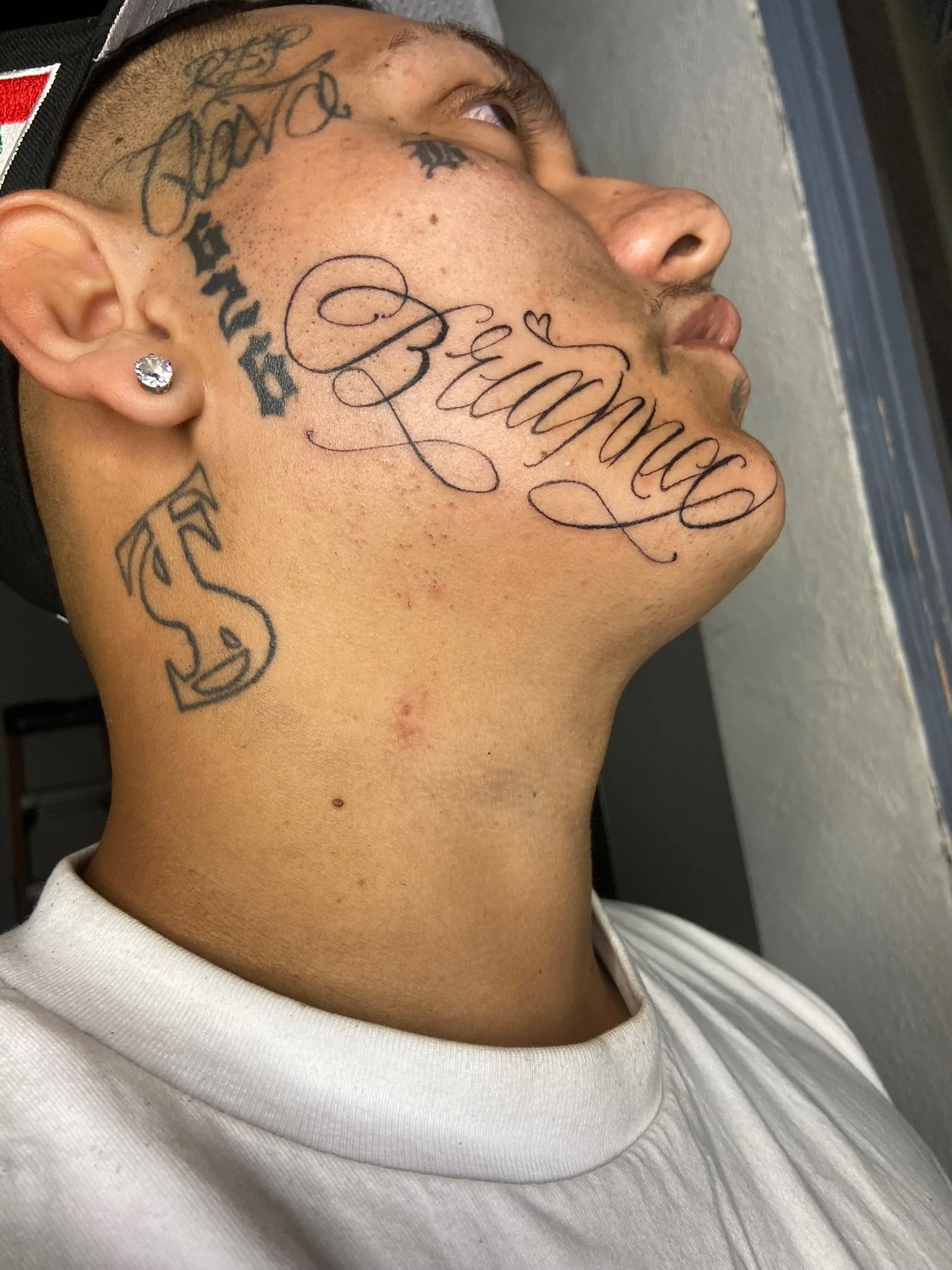 Close-up of a person's side face and neck with tattoos, including the name 'Benjamin' in cursive with a small heart, and other tattoos on the ear, cheek, and neck. The person is wearing a white shirt and has a diamond stud earring.