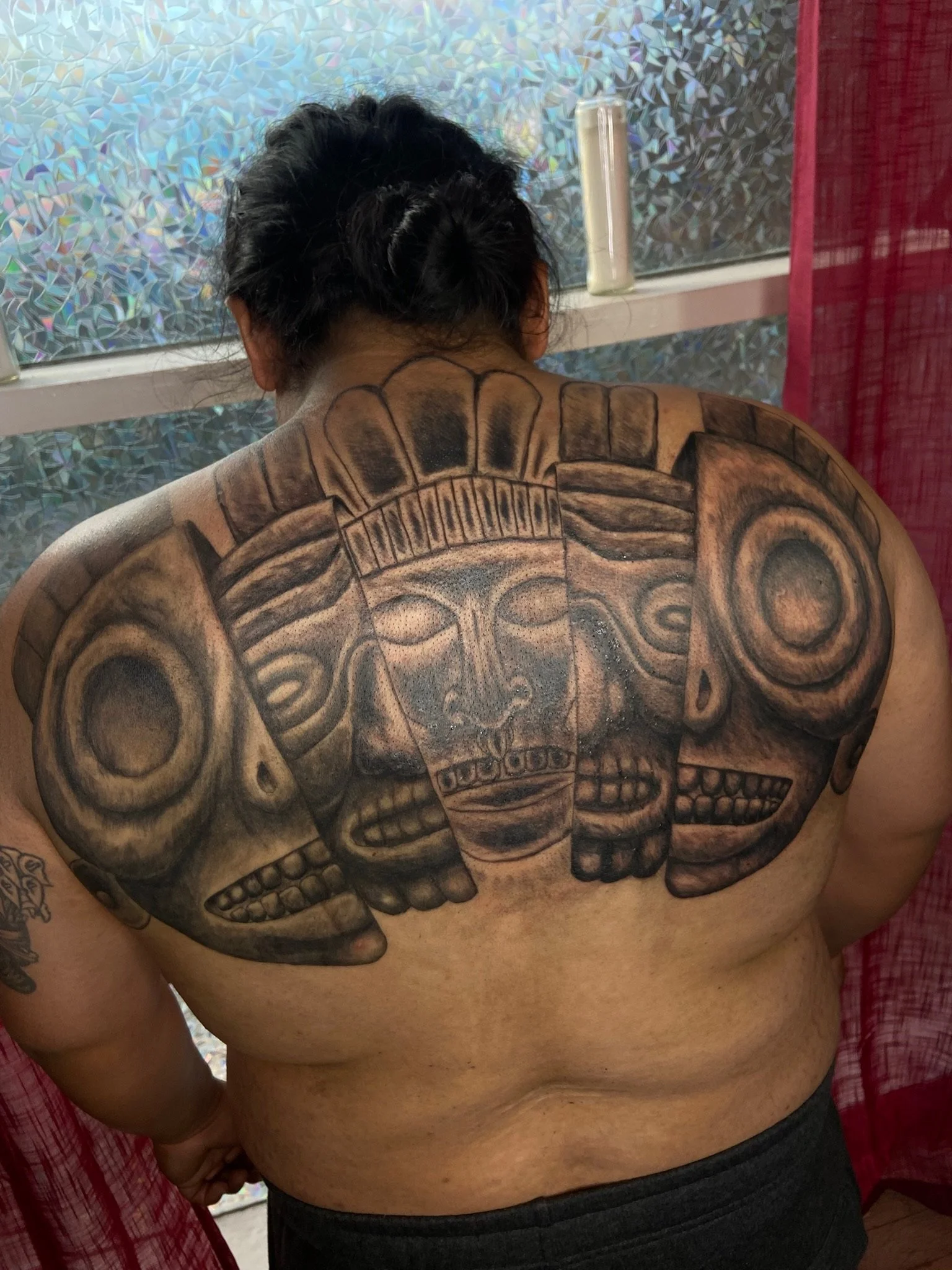 A person with a large tattoo of a skull on their back, viewed from behind. The person has short, dark hair and is shirtless. The background includes a frosted window and a red curtain.