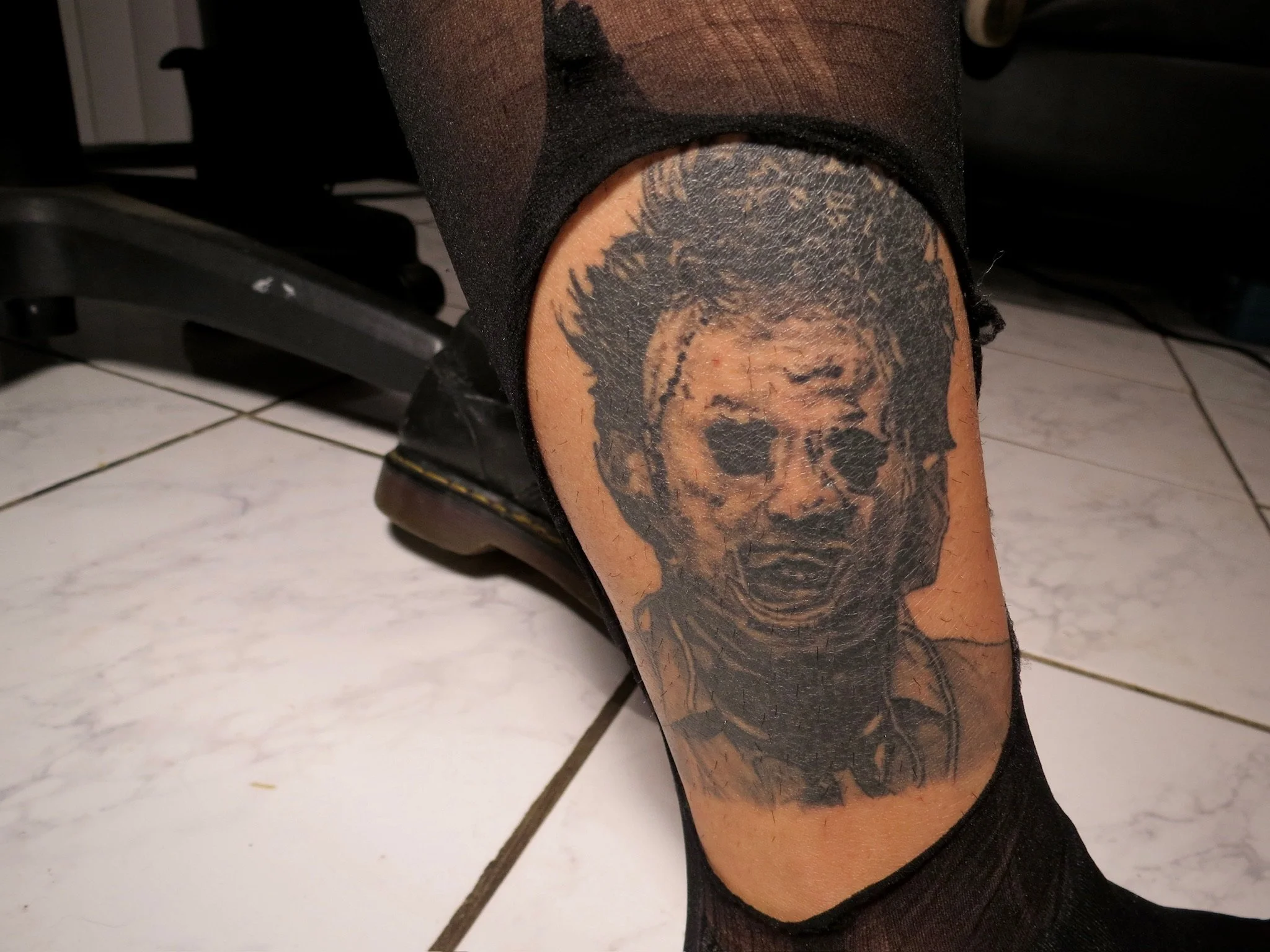 Close-up of a person's leg with a large tattoo of a skull on the thigh, wearing black shorts and shoes, next to a treadmill on a tiled floor.