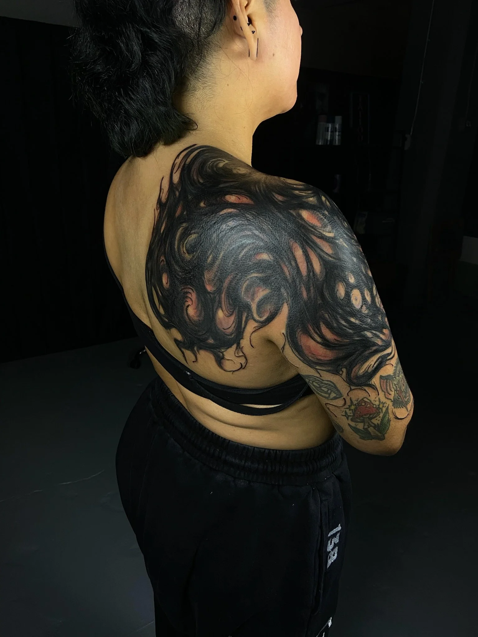 A person with short dark hair showing a large blackwork swirl tattoo that is a large scale coverup on their shoulder and upper arm, wearing black shorts with white text and black pants, standing against a dark background.