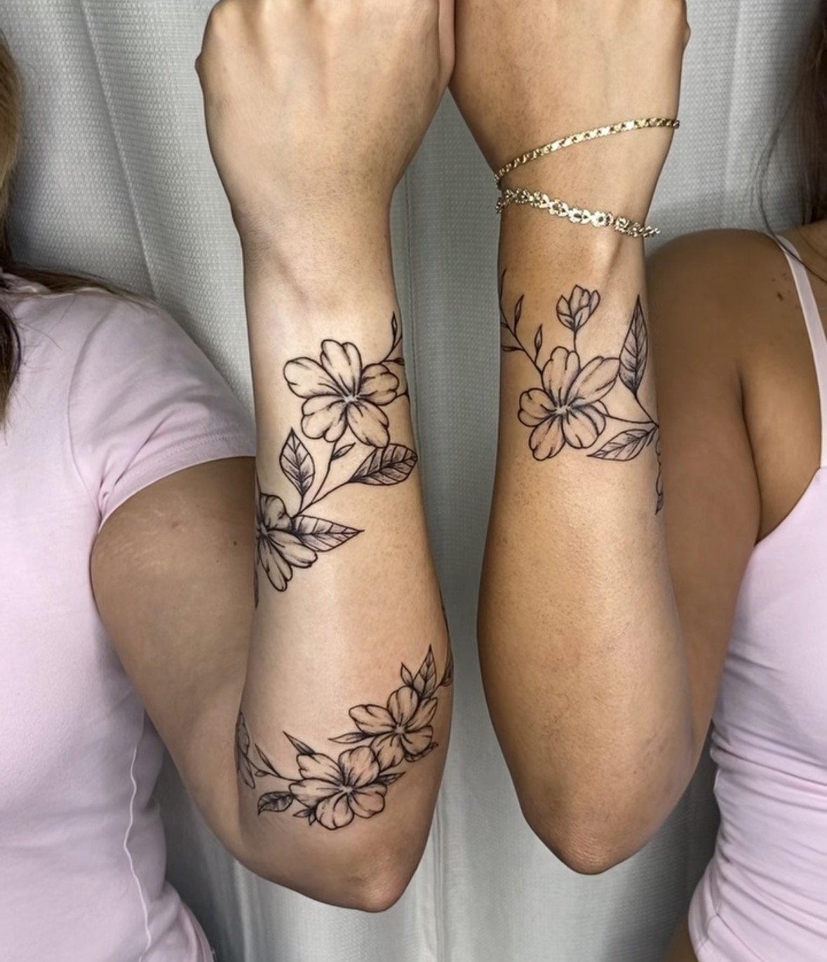 Two women lying down with their arms raised, displaying matching floral tattoos on their forearms and upper arms. One woman has a bracelet on her wrist. Matching tattoos by artist Agua at Floating Streams Tattoo.