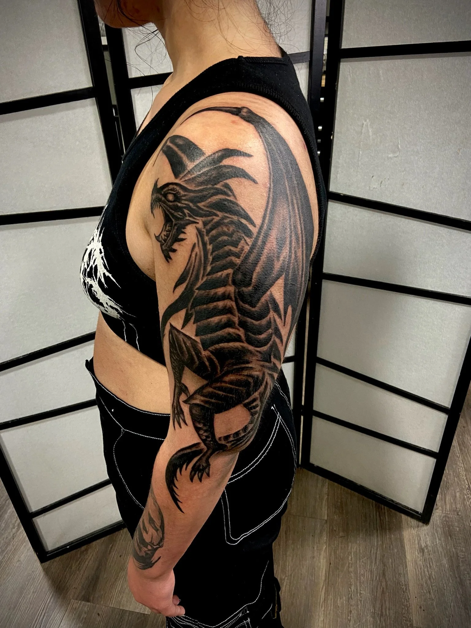 A woman with a large black dragon tattoo on her upper arm and forearm, wearing a black sleeveless shirt, standing in front of a folding room divider with a wooden floor.