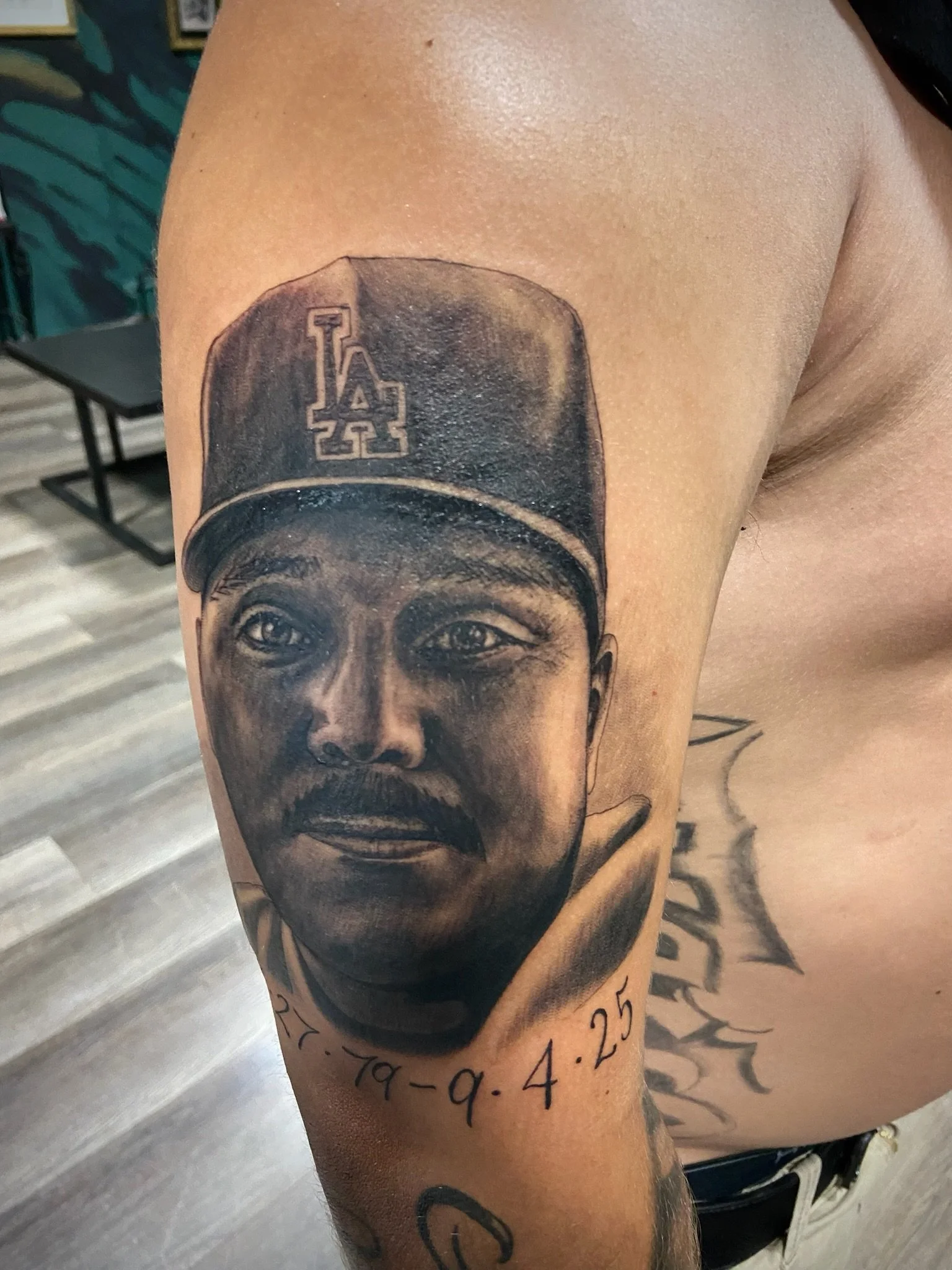 Tattoo of a realistic black and gray portrait of a child's face wearing a Los Angeles Dodgers baseball cap on a person's upper arm. Below the portrait, there are dates written in black ink.