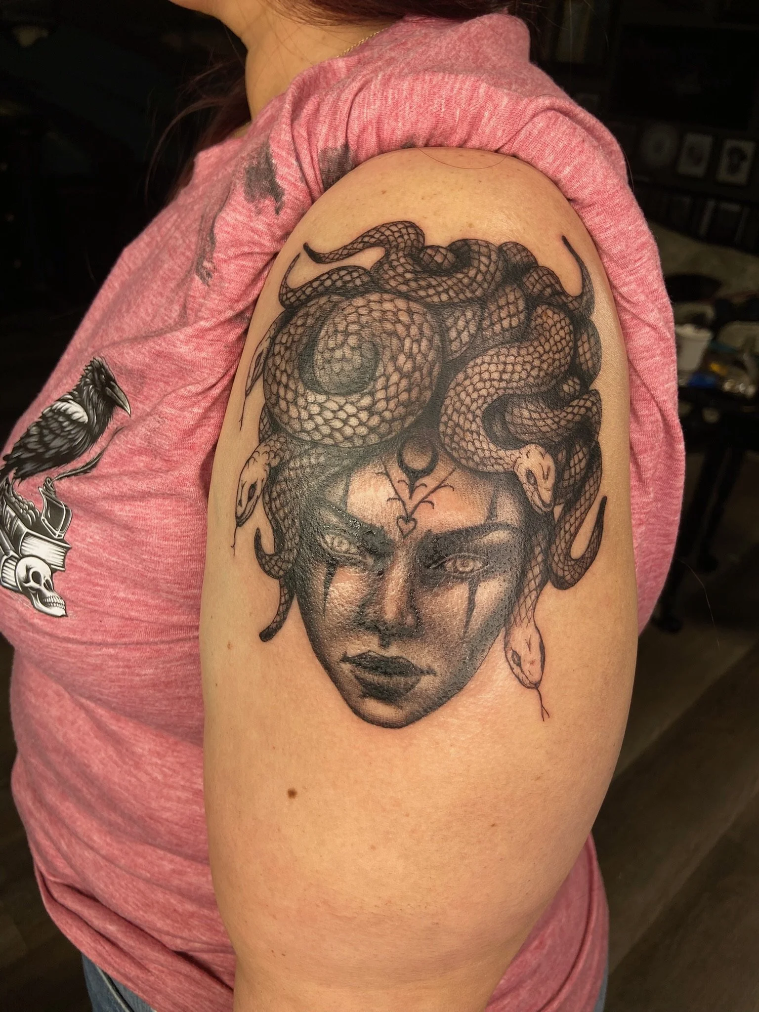 A black and gray tattoo on a person's upper arm, depicting a woman's face with bold makeup, with a snake intertwined around her head and face.