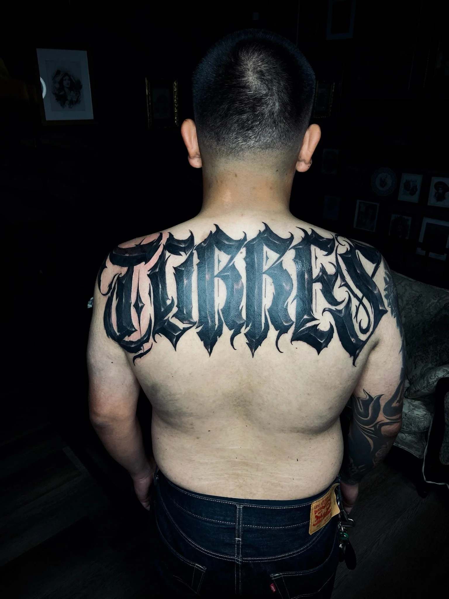 Back of a person with short black hair, shirtless, showing a large tattoo across the upper back and shoulders, with dark ink and bold lettering.