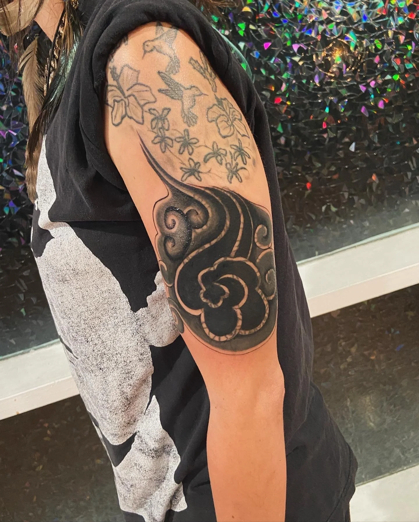A person's upper arm with a large tattoo consisting of black and gray designs, including a tree branch, flowers, and a bird, against a black background.