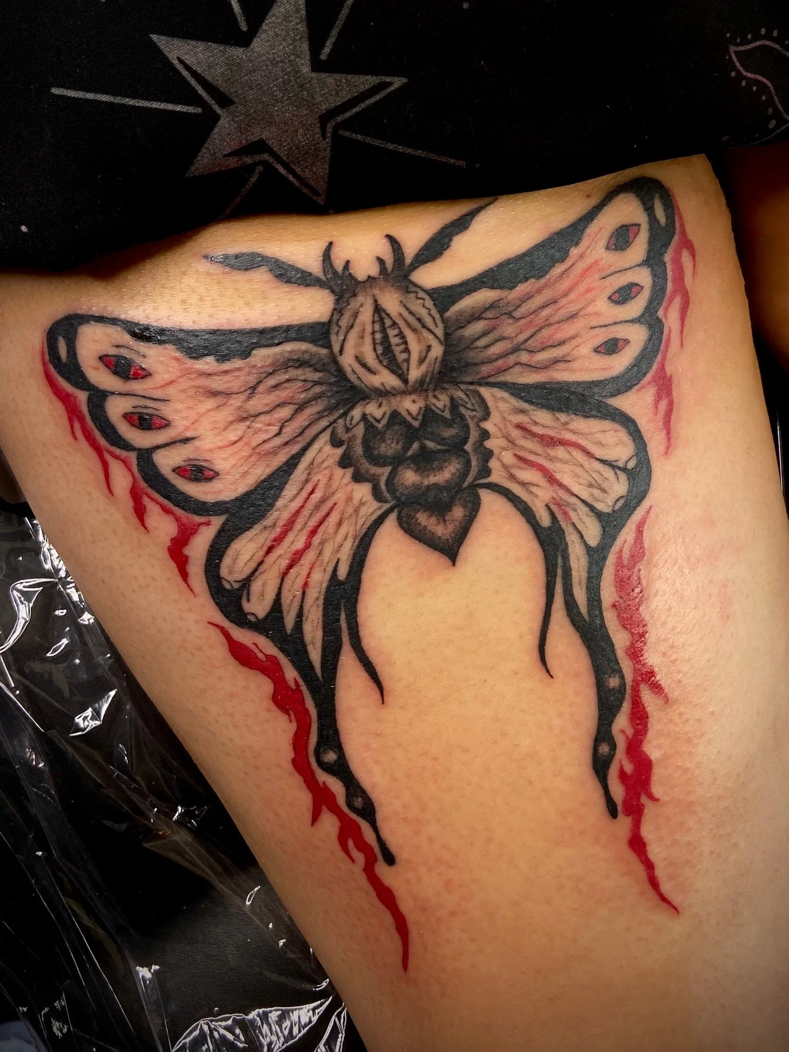 New tattoo of a butterfly with black outlines and red accents, surrounded by red ink resembling blood, on a person's skin.