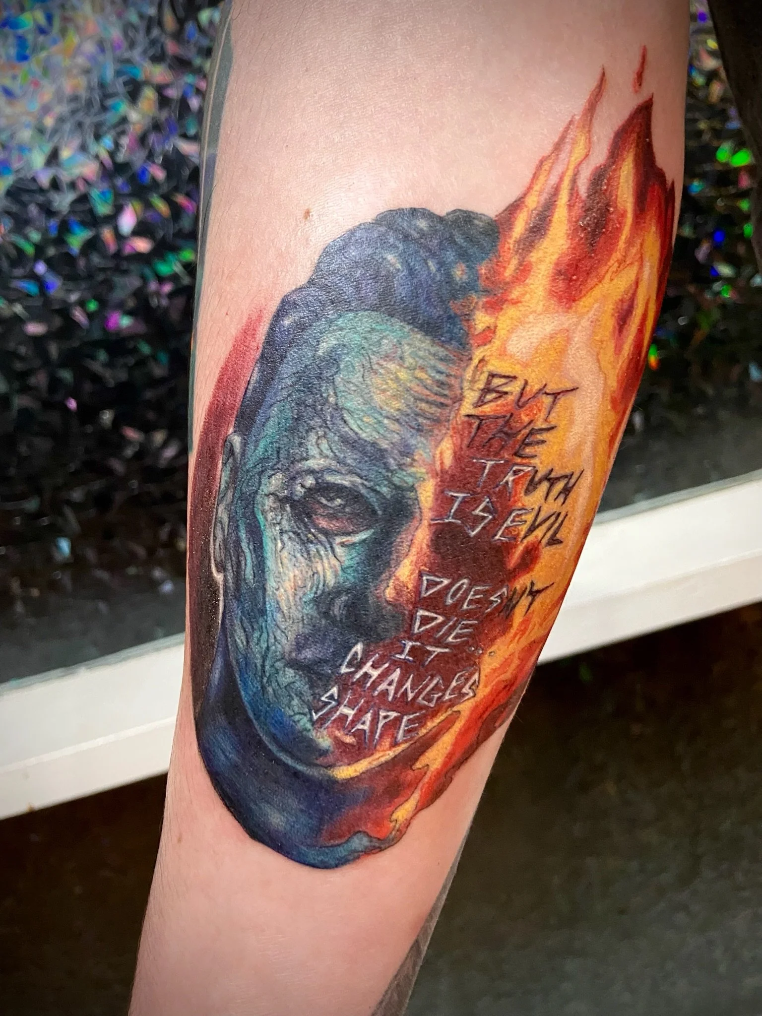 A realistic-style tattoo on a person's arm depicts a human skull and a fiery explosion with the words 'but the truth is I feel death' written across it.