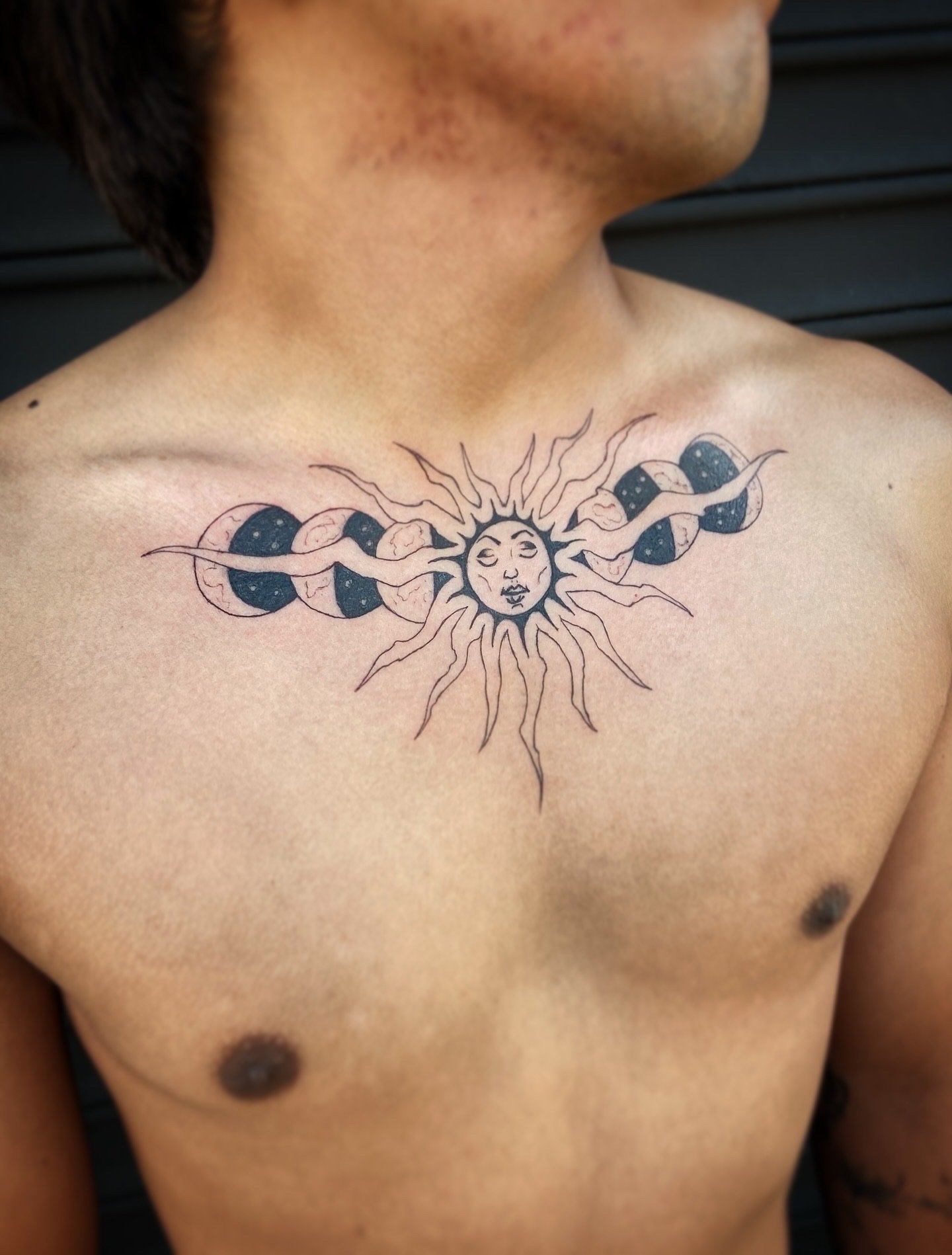 Tattoo of a sun with a human face and rays, surrounded by phases of the moon, on a person's chest.