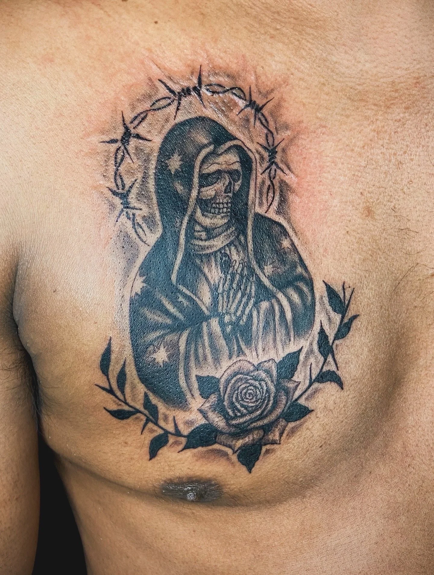 Tattoo of a skeleton wrapped in a hooded cloak, surrounded by a circular thorny vine and a rose at the bottom.