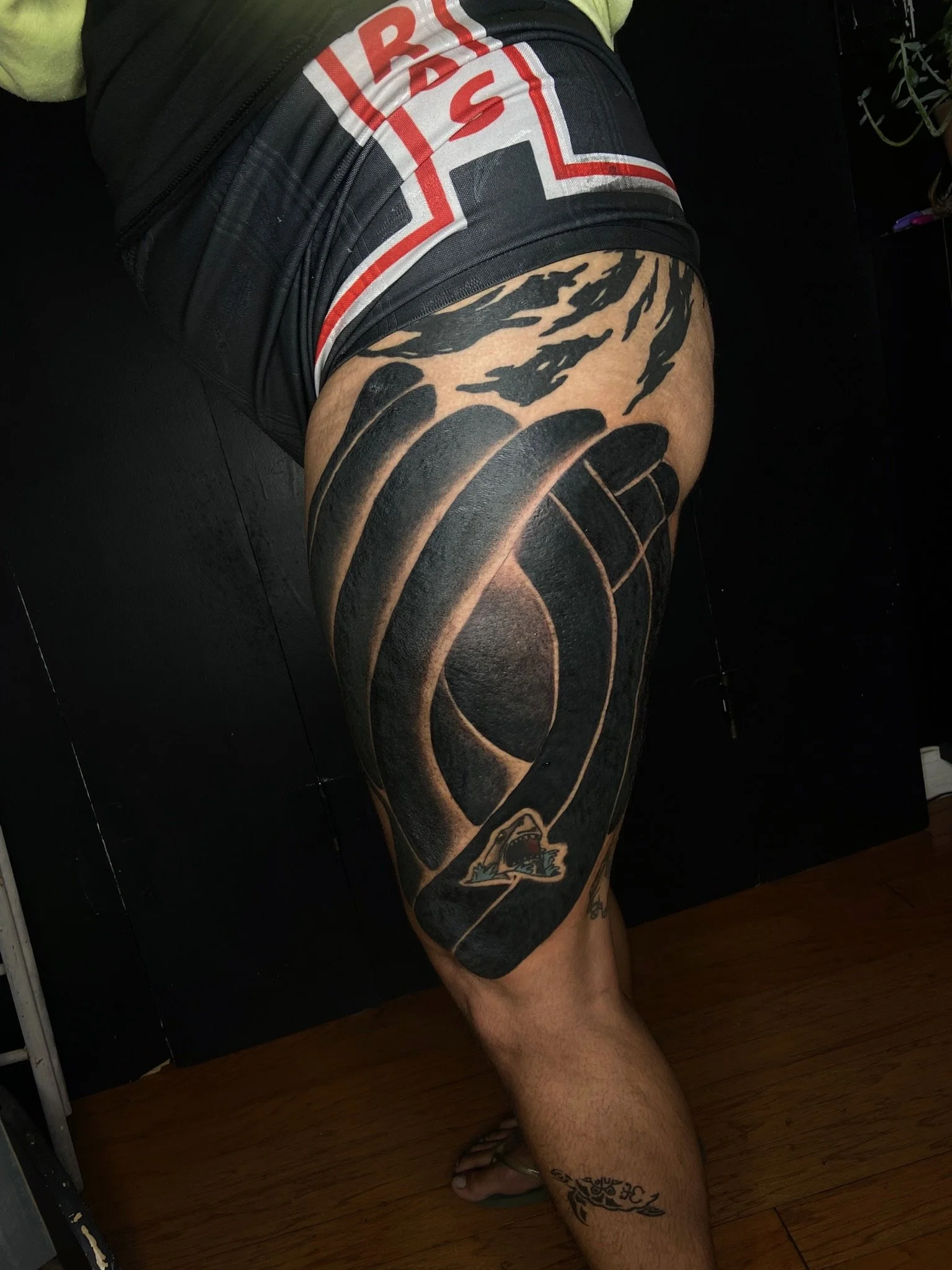 A person's lower body, lying on a dark surface, wearing black shorts with red and white lettering, and sporting tattoos on their thigh and calf. The tattoo on the thigh resembles a wolf or similar animal, and the calf tattoo is a decorative tribal or