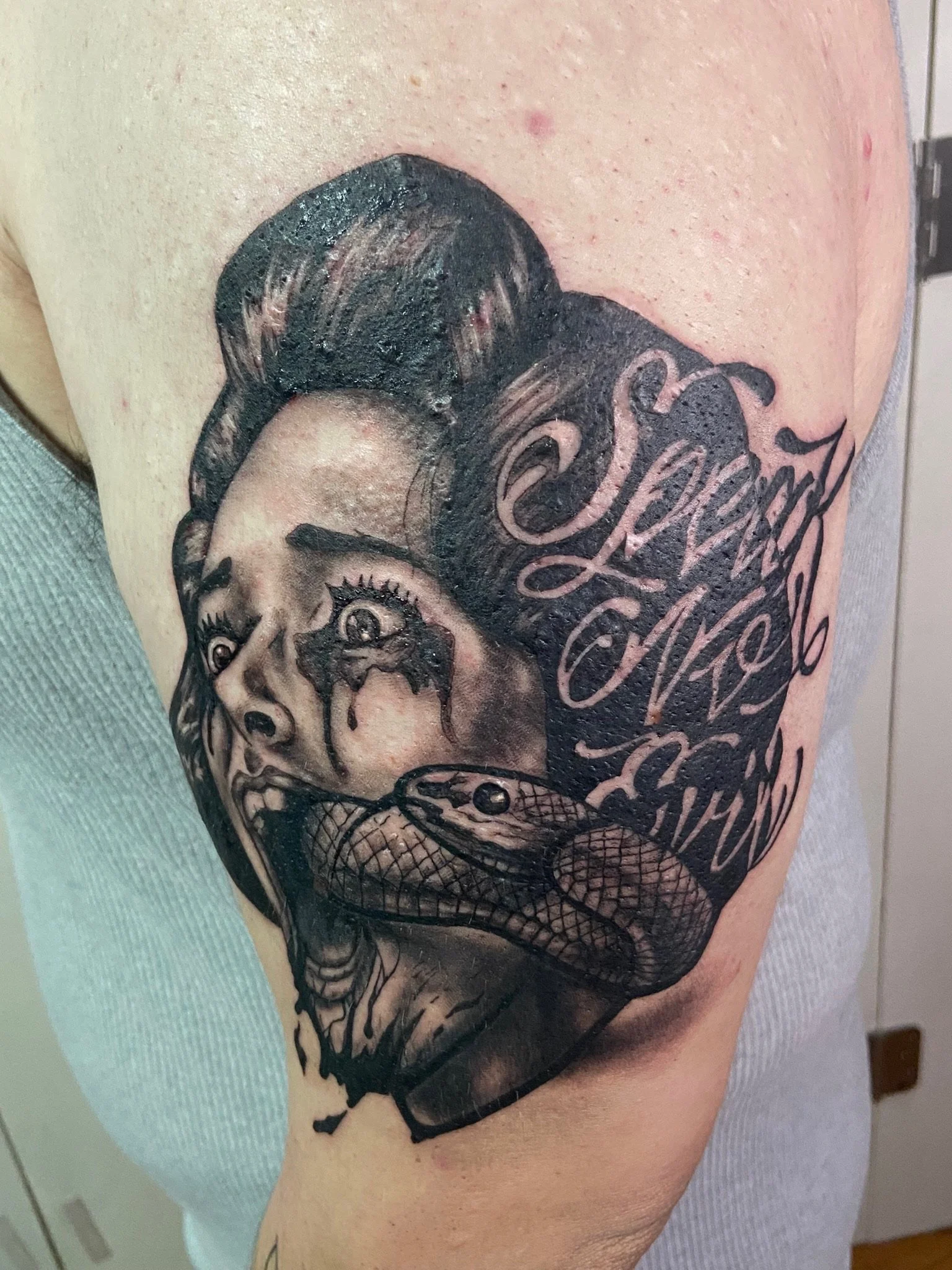 Tattoo of a woman's face with makeup on her eyes and lips, black and gray shading, with ornate, decorative text surrounding and partly overlapping her face.