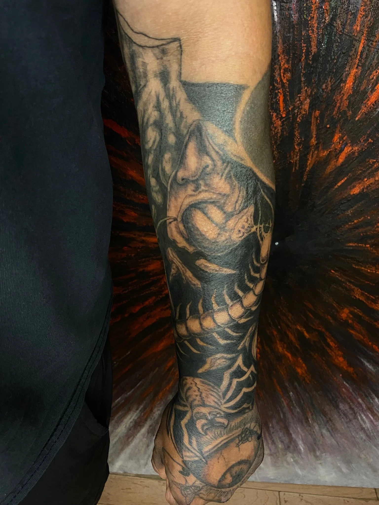 Close-up of a person's arm with detailed black and gray tattoos, including a hydra-like creature, a snake, and various abstract designs, against a dark, fiery background.