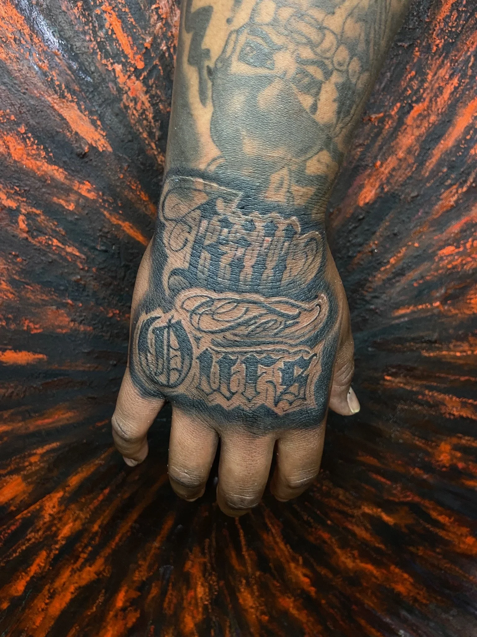 Close-up photo of a person's hand with tattoos against a black and orange background, with the focus on the tattooed hand.