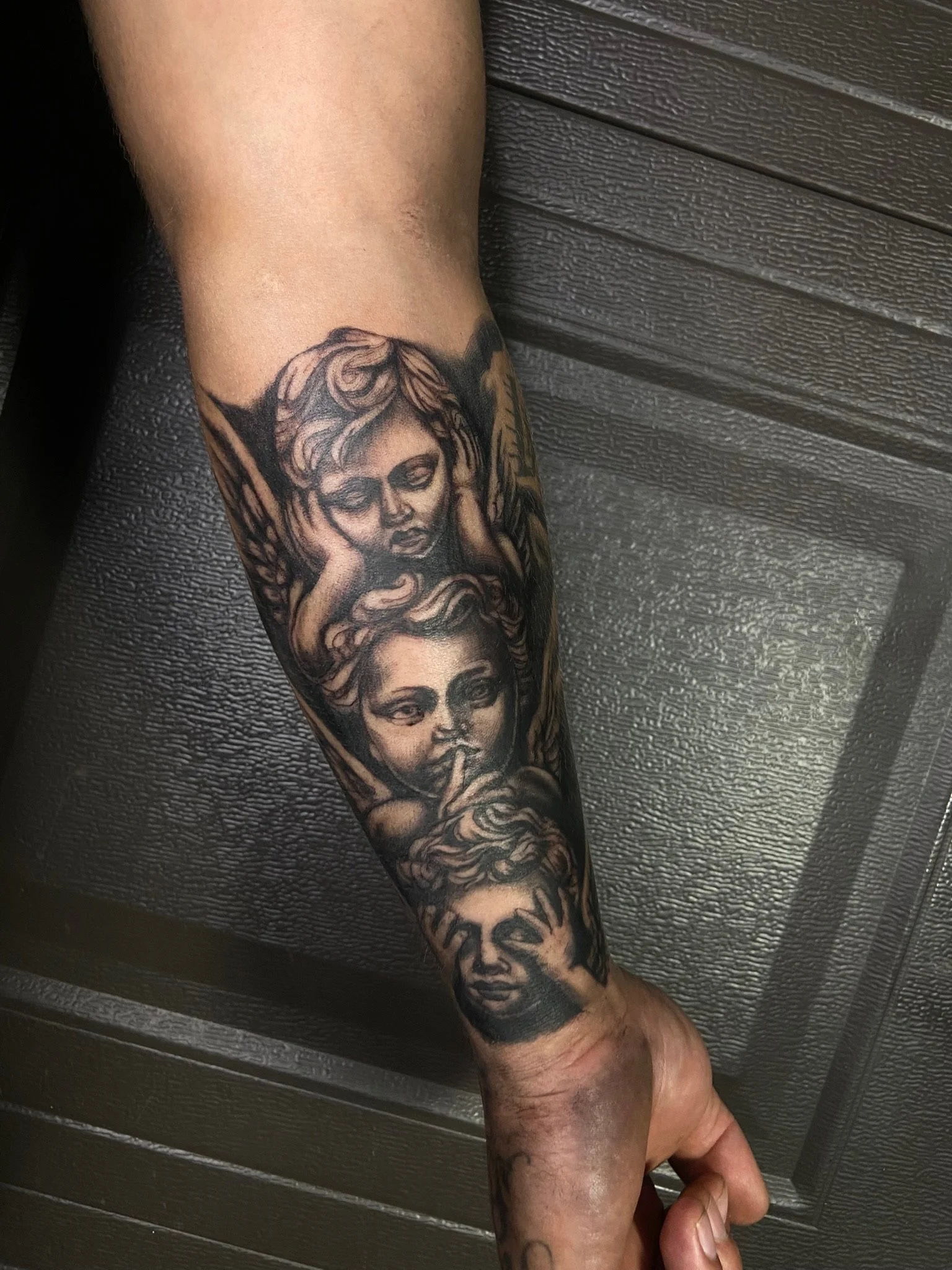 A tattooed arm with various black and gray tattoos, including faces, holding a penis, against a dark textured surface background.