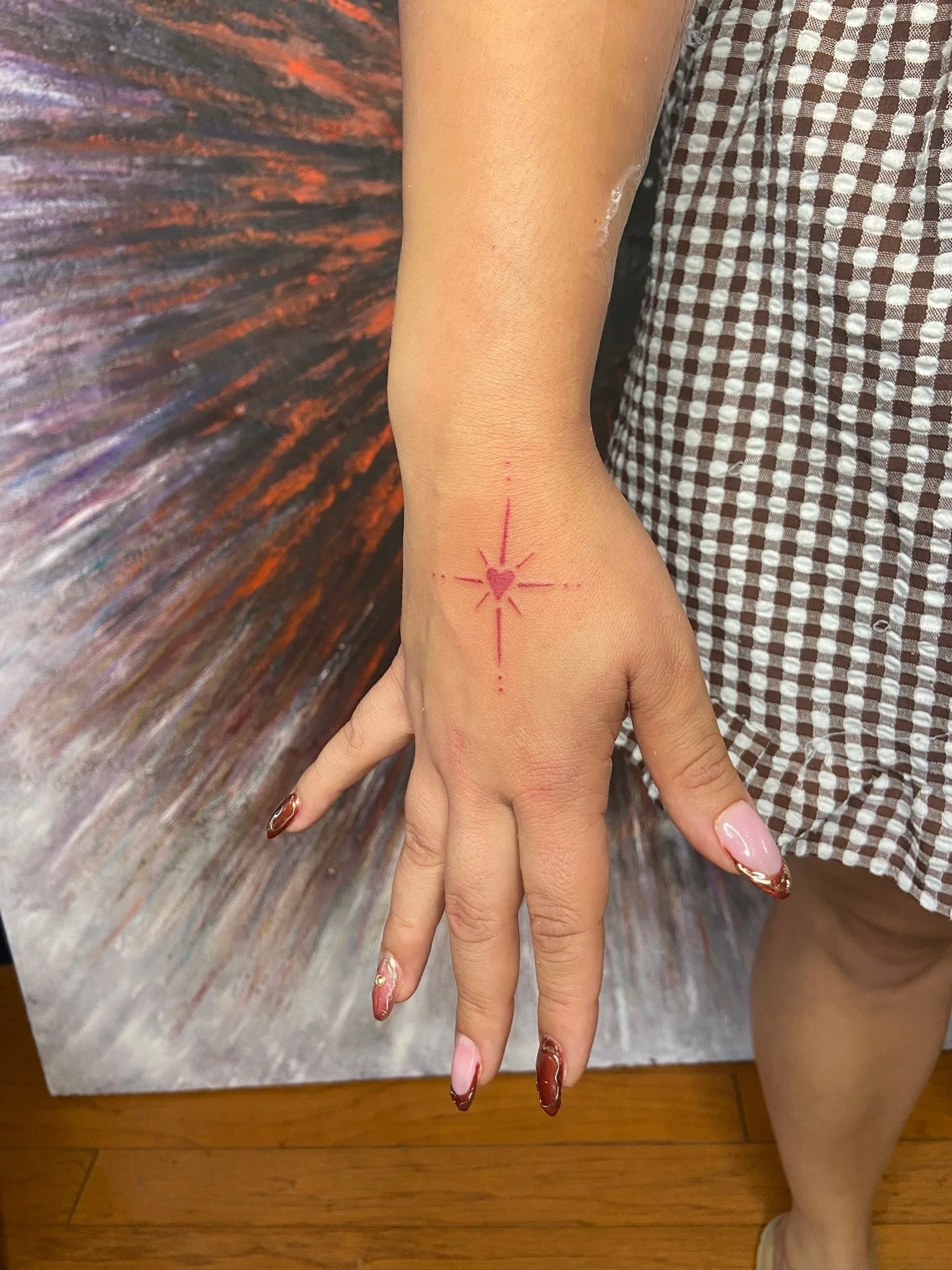 A person's hand resting on a surface with a colorful, textured background. The hand has decorative nail polish and a small tattoo of a heart with radiating lines and dots on the wrist.