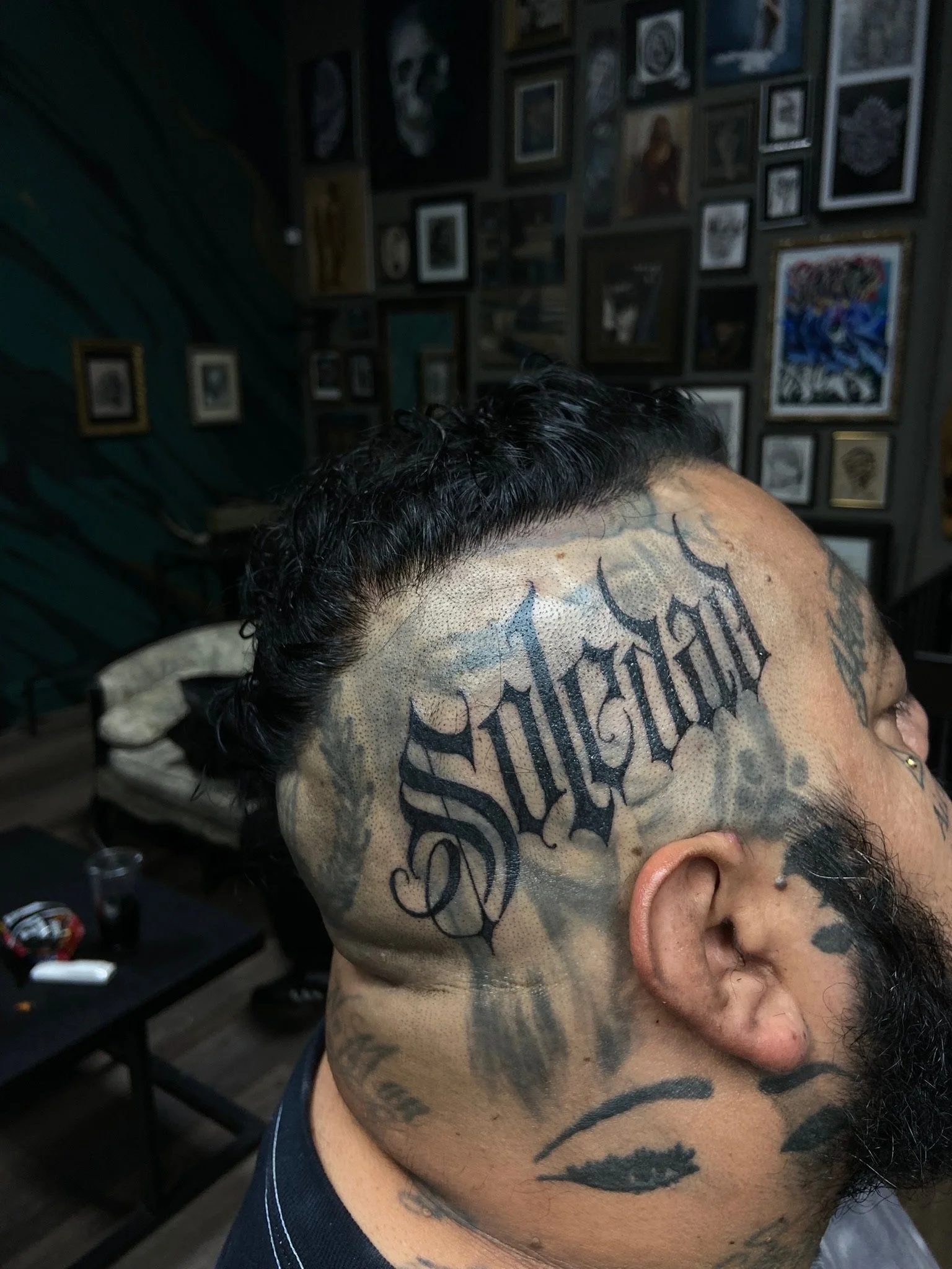 A person with short, dark, curly hair tattoos and a fade haircut, with a large tattoo on the side of their head that spells out 'Respect' in gothic font, inside a tattoo studio with numerous framed pictures on the wall.