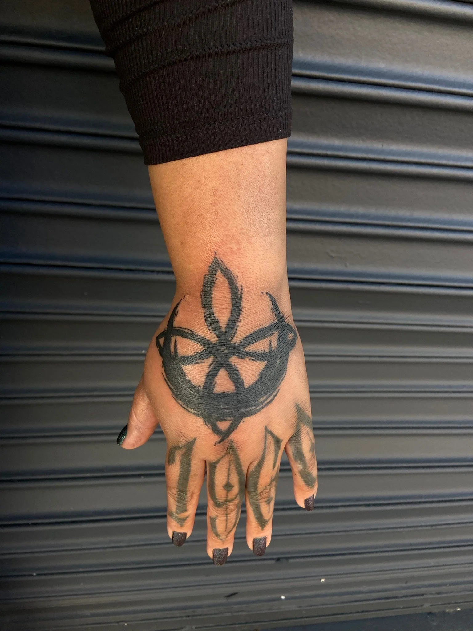 A tattooed hand with black nail polish in front of a metal roller shutter background.