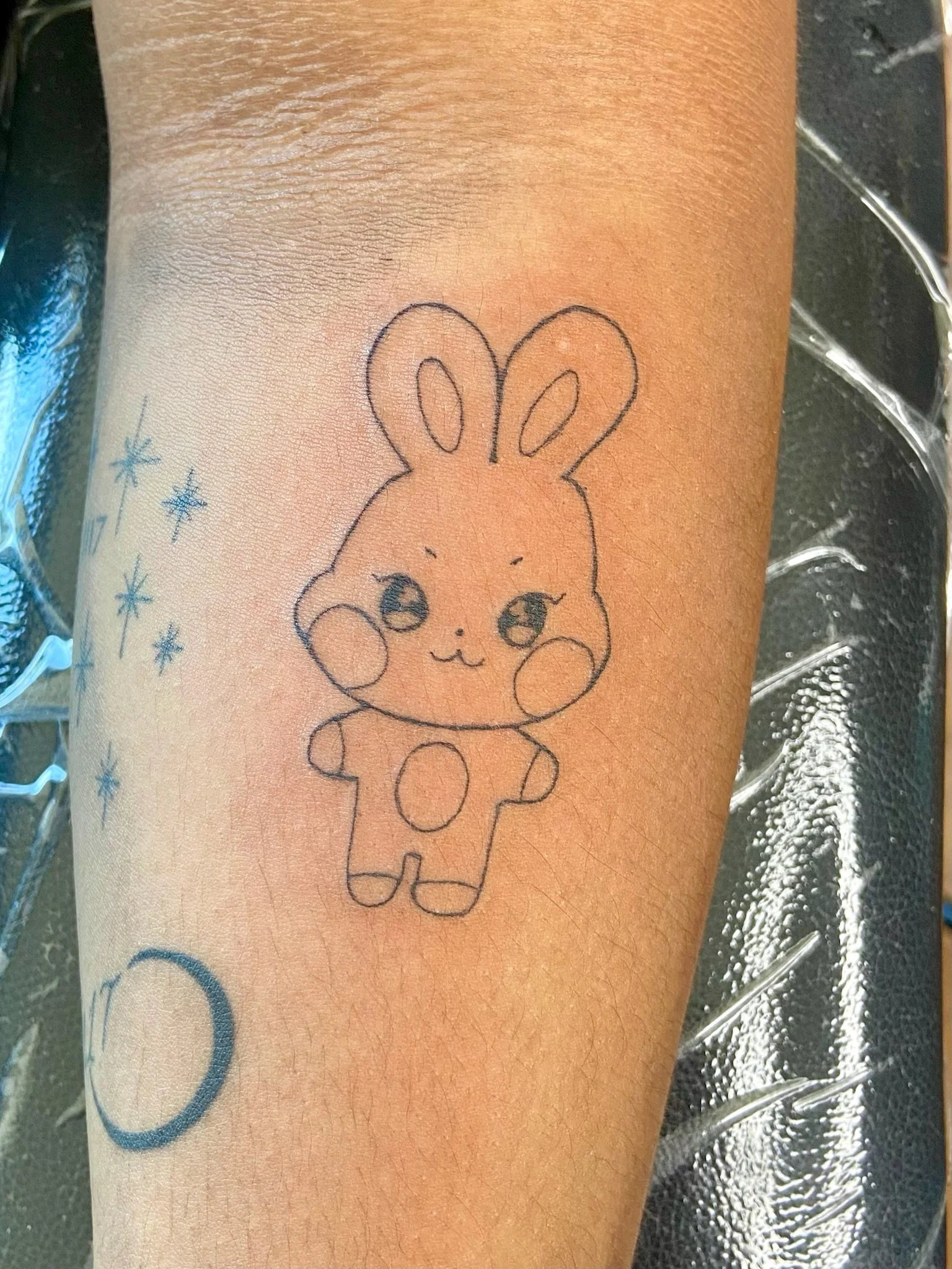 Tattoo of a cute bunny character with large ears, on a person's arm, surrounded by small stars and symbols.