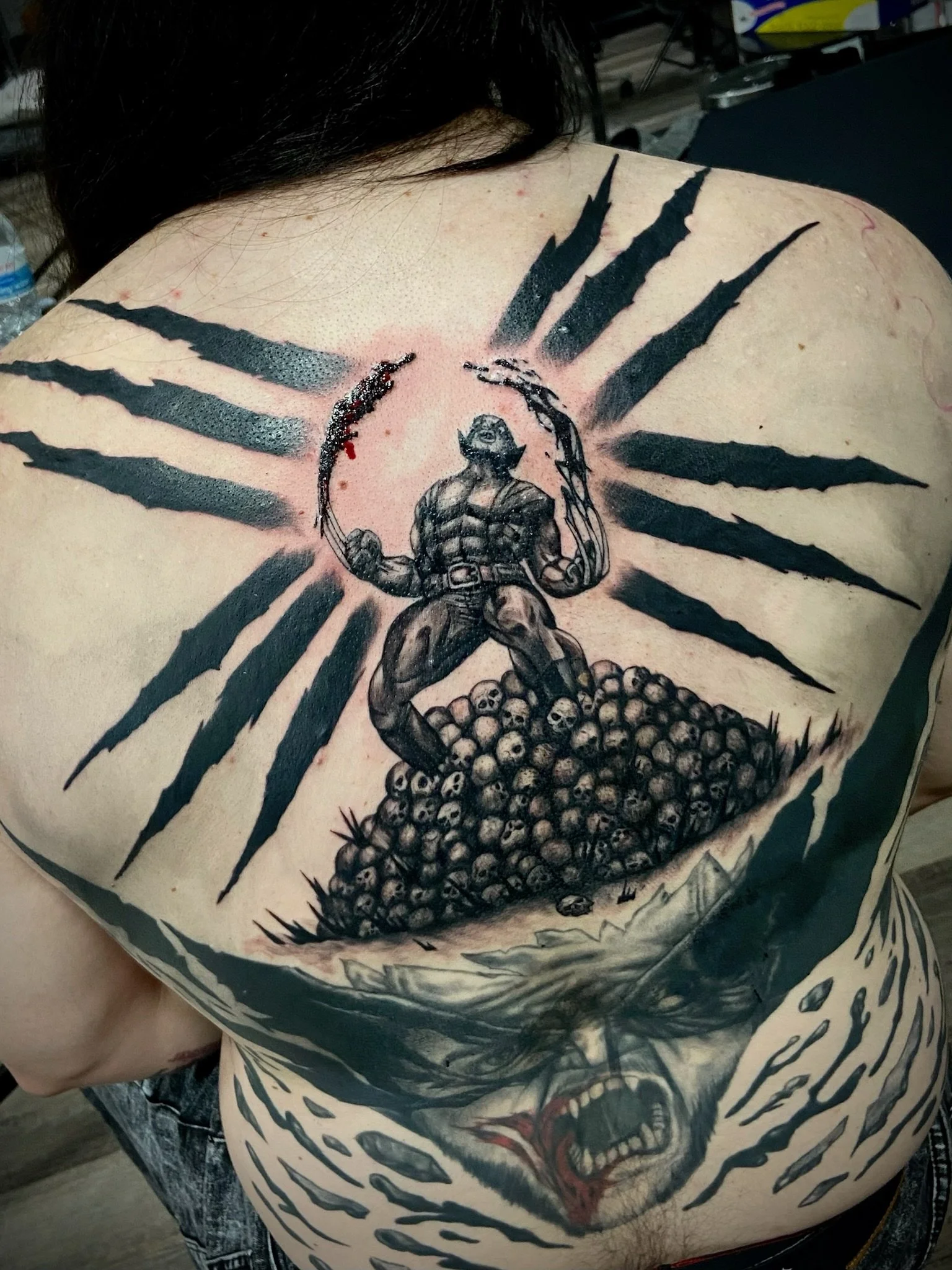 A person's back with a new tattoo of a muscular man holding a sword, surrounded by stylized black claw marks, with a pile of skulls at the bottom.