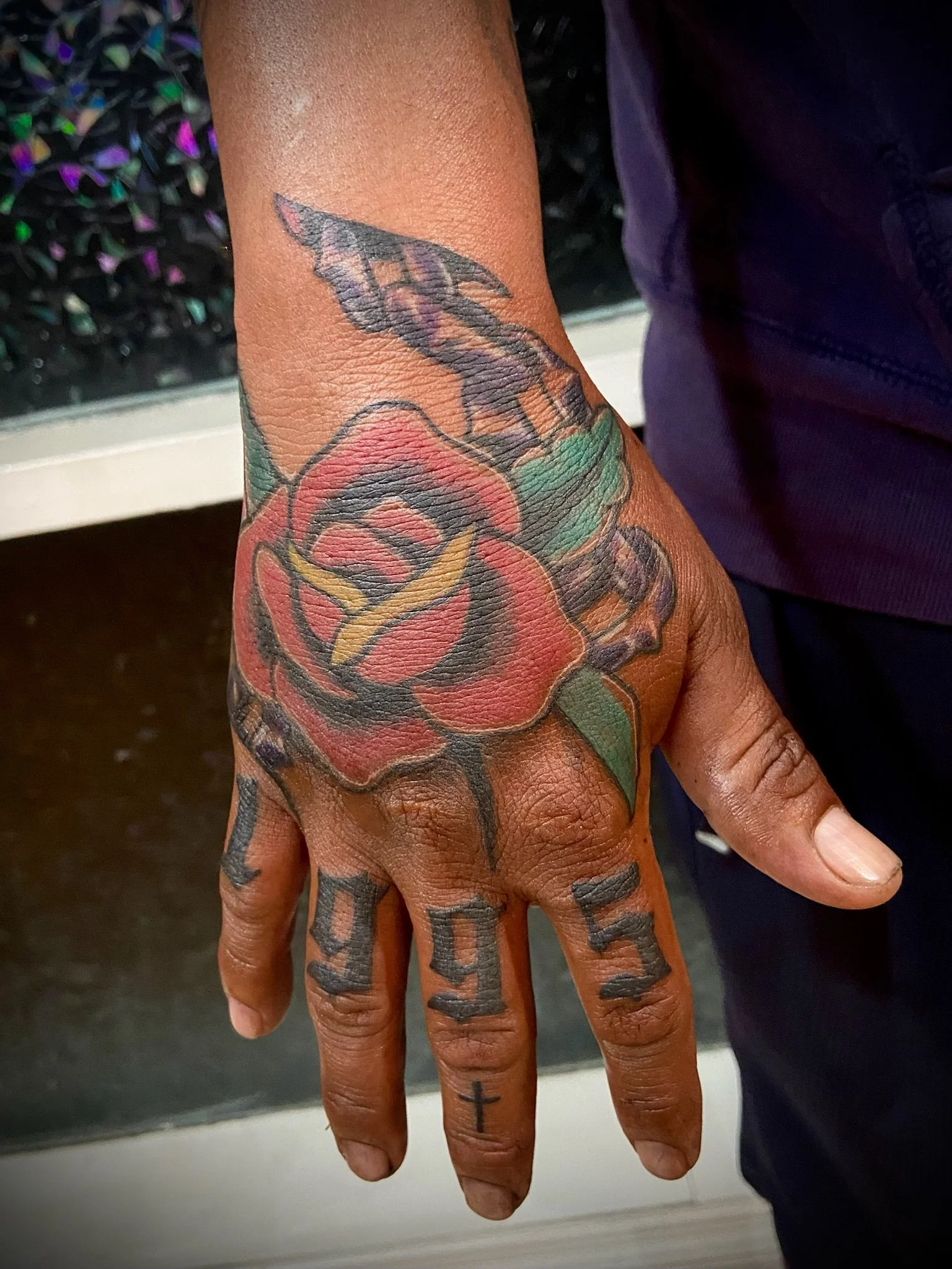 Close-up of a person's hand with tattoos, including a cross tattoo on the finger, and a colorful flower tattoo on the wrist.