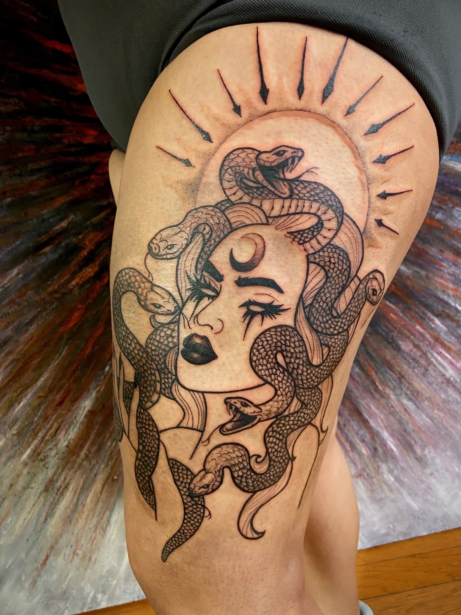 A person's thigh with a tattoo of a snake intertwined with a woman's face, surrounded by a semi-circular clock or sunburst design.