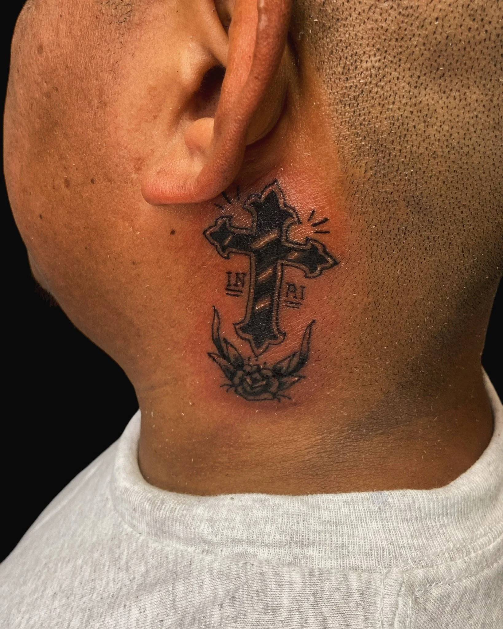 A close-up of a person's face showing nose and lips, with a tattoo of a cross on the side of the neck, and wearing a white shirt.