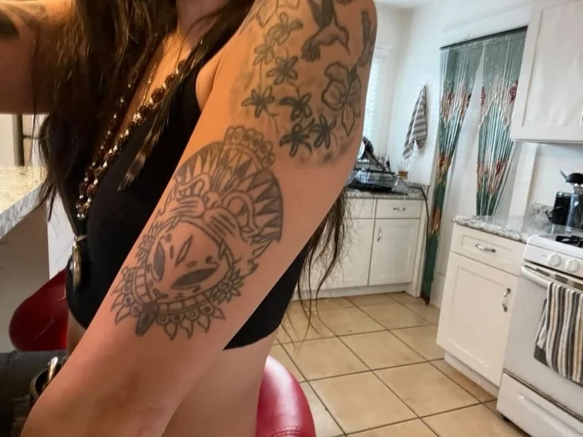 Close-up of a woman's arm with tattoos, including a large design of a cow's face surrounded by decorative elements, on a black sleeveless top in a kitchen setting.