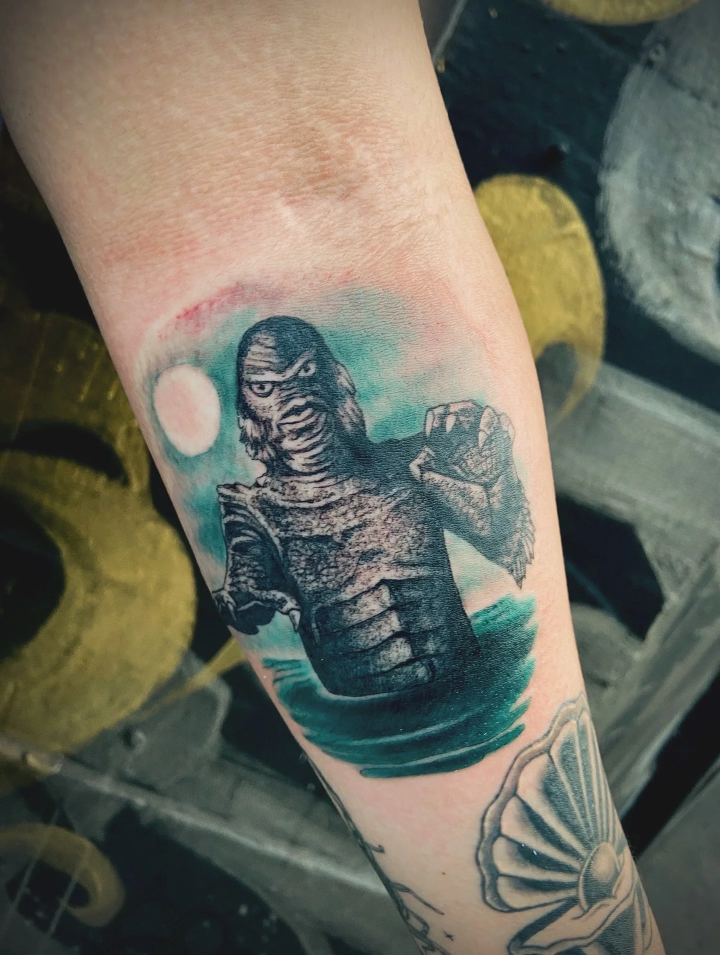 Tattoo of a fierce-looking warrior or superhero with a face mask on a person's upper arm, with the background featuring a light blue-greenish color and a setting sun.