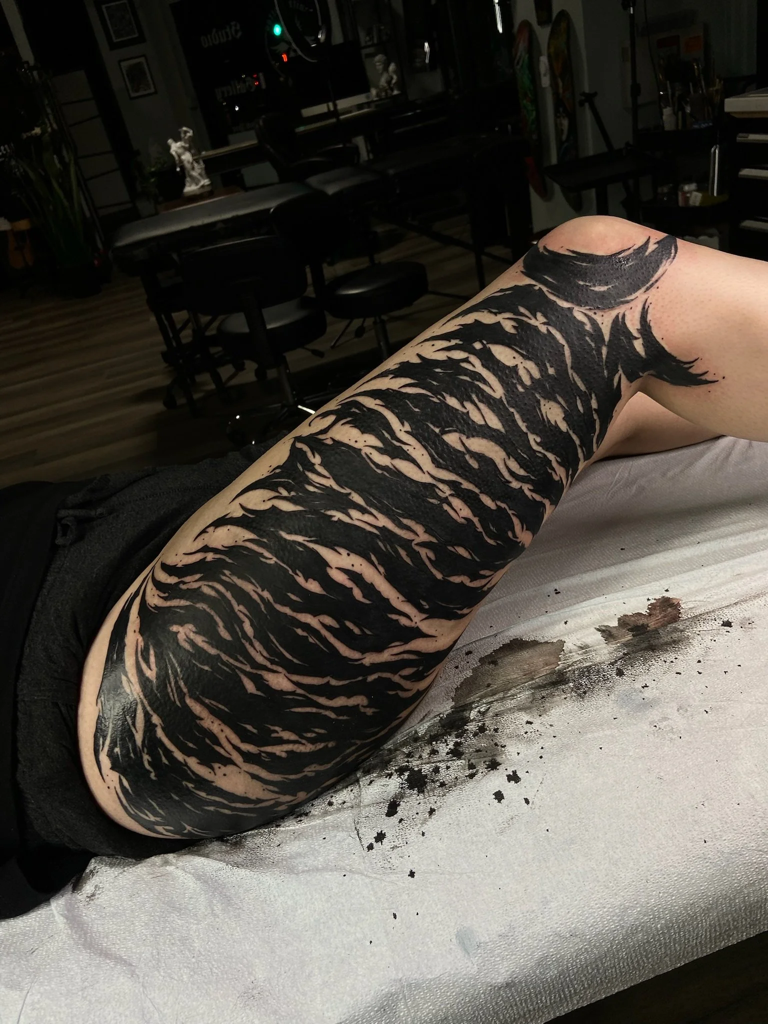A person's arm with a black abstract tattoo resting on a surface, with black ink splatters and spray paint around. The background appears to be a dimly lit room with chairs, tables, and artwork on the walls.